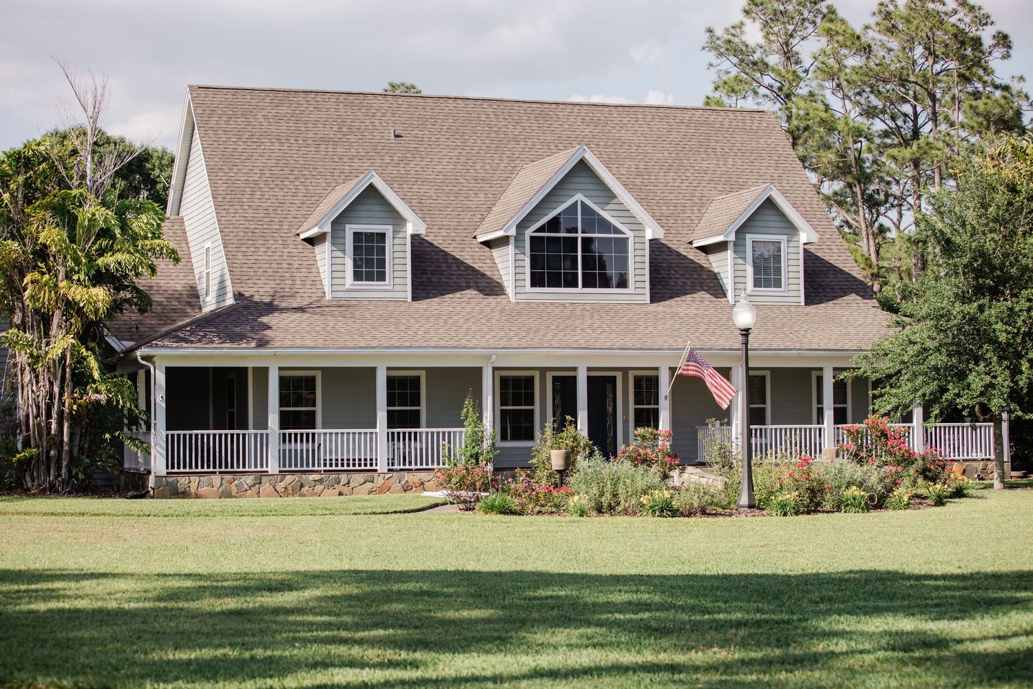 A beautiful Cape Cod style home with a wrap-around porch and manicured lawn displaying an American flag.