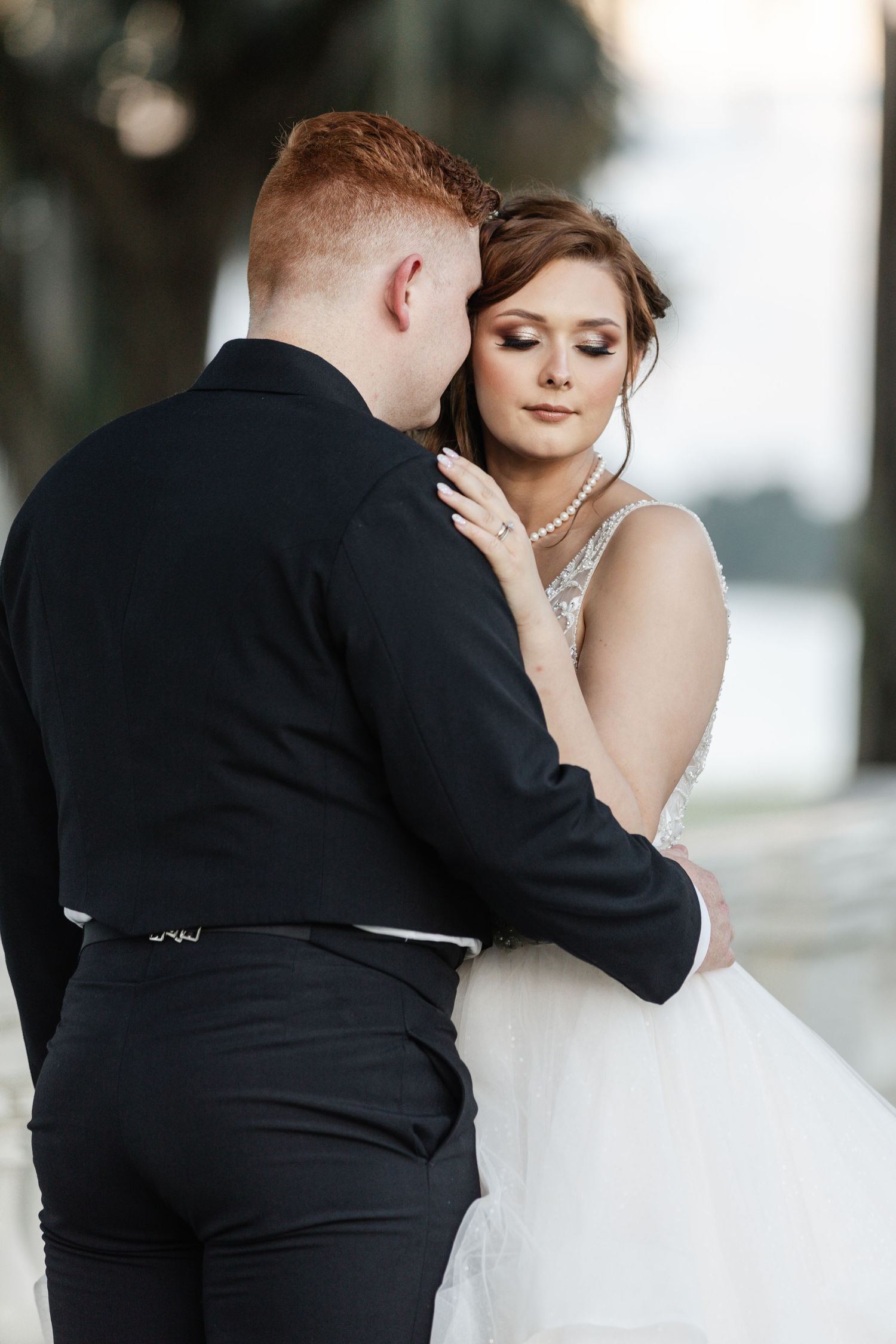 Bride in a beaded white gown embracing a red-haired groom in a black suit outdoors.