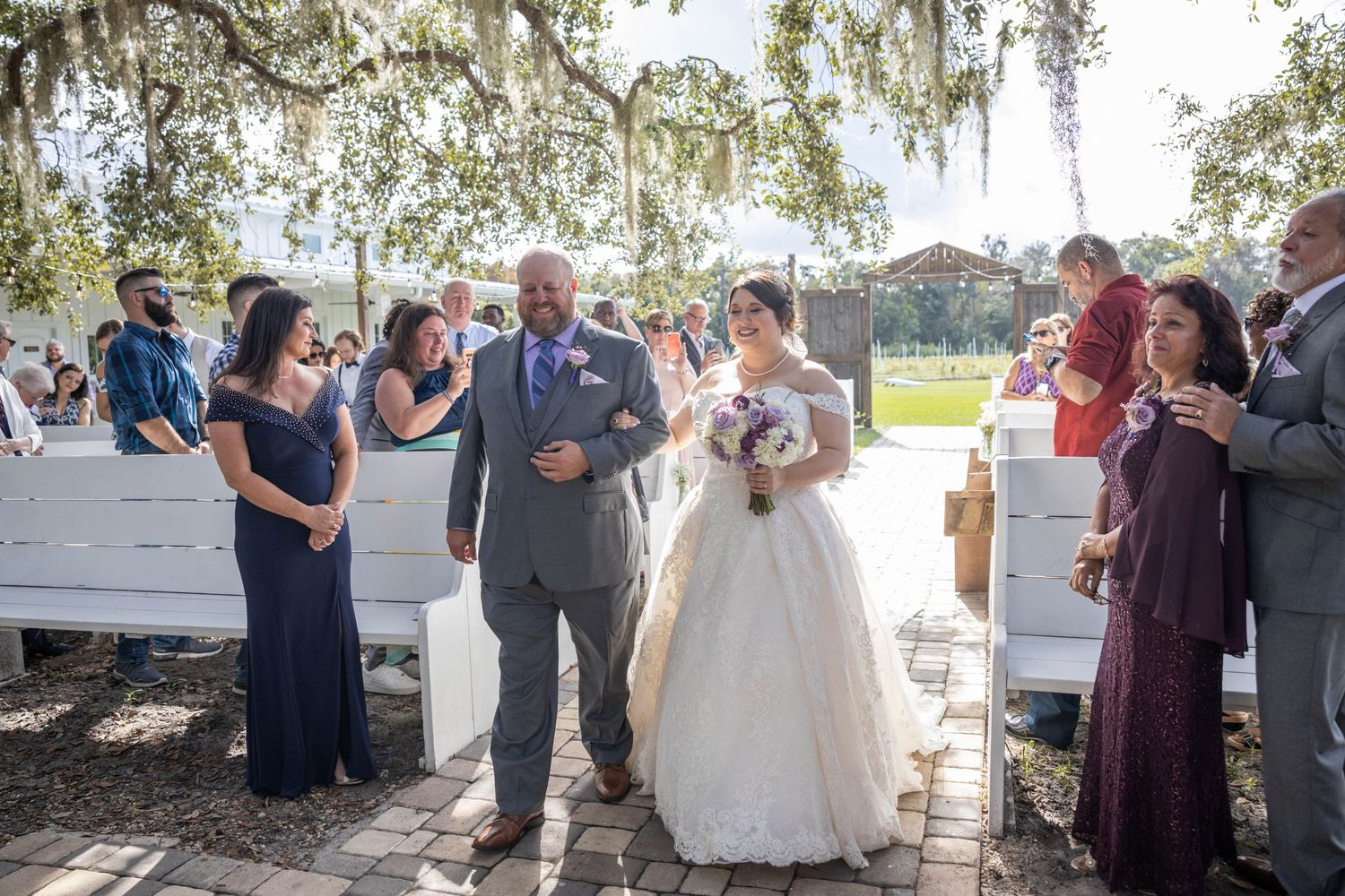 Bride in white lace gown walks down outdoor aisle with escort past seated guests under Spanish moss trees.