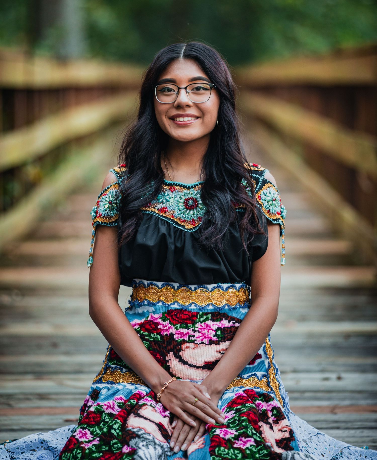 Young woman in colorful embroidered traditional Mexican dress sits smiling on a wooden bridge outdoors.