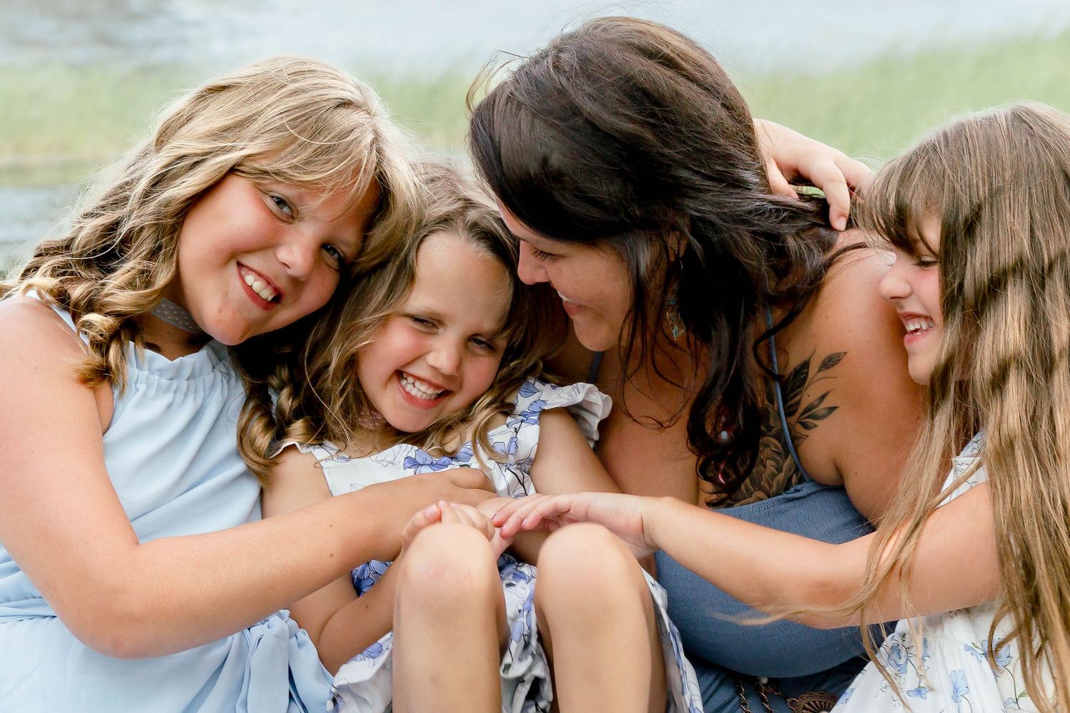 A joyful mother laughs and cuddles with her three smiling daughters by the water in blue dresses.