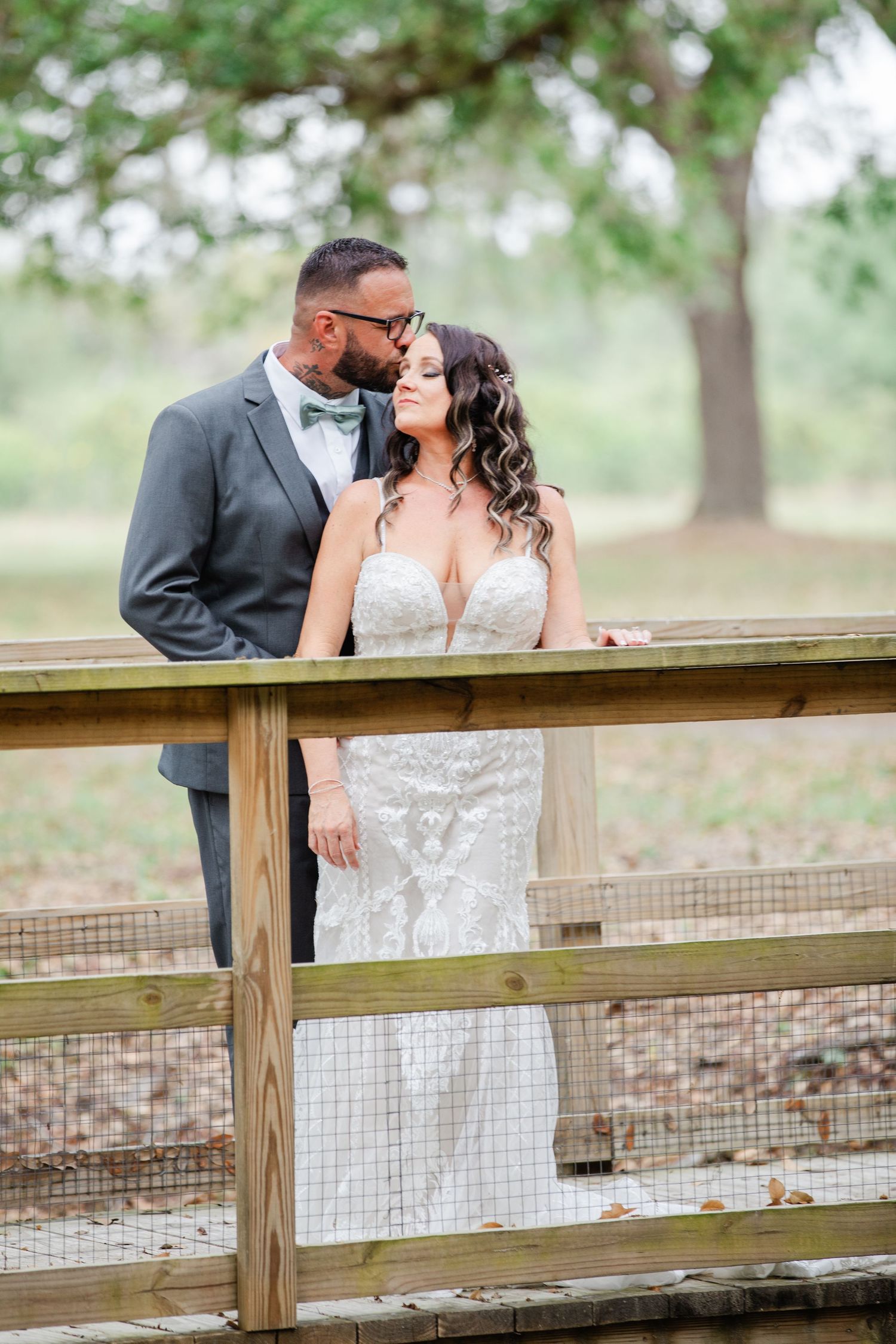 Groom in gray suit kisses bride on forehead near rustic wooden fence at outdoor wedding.