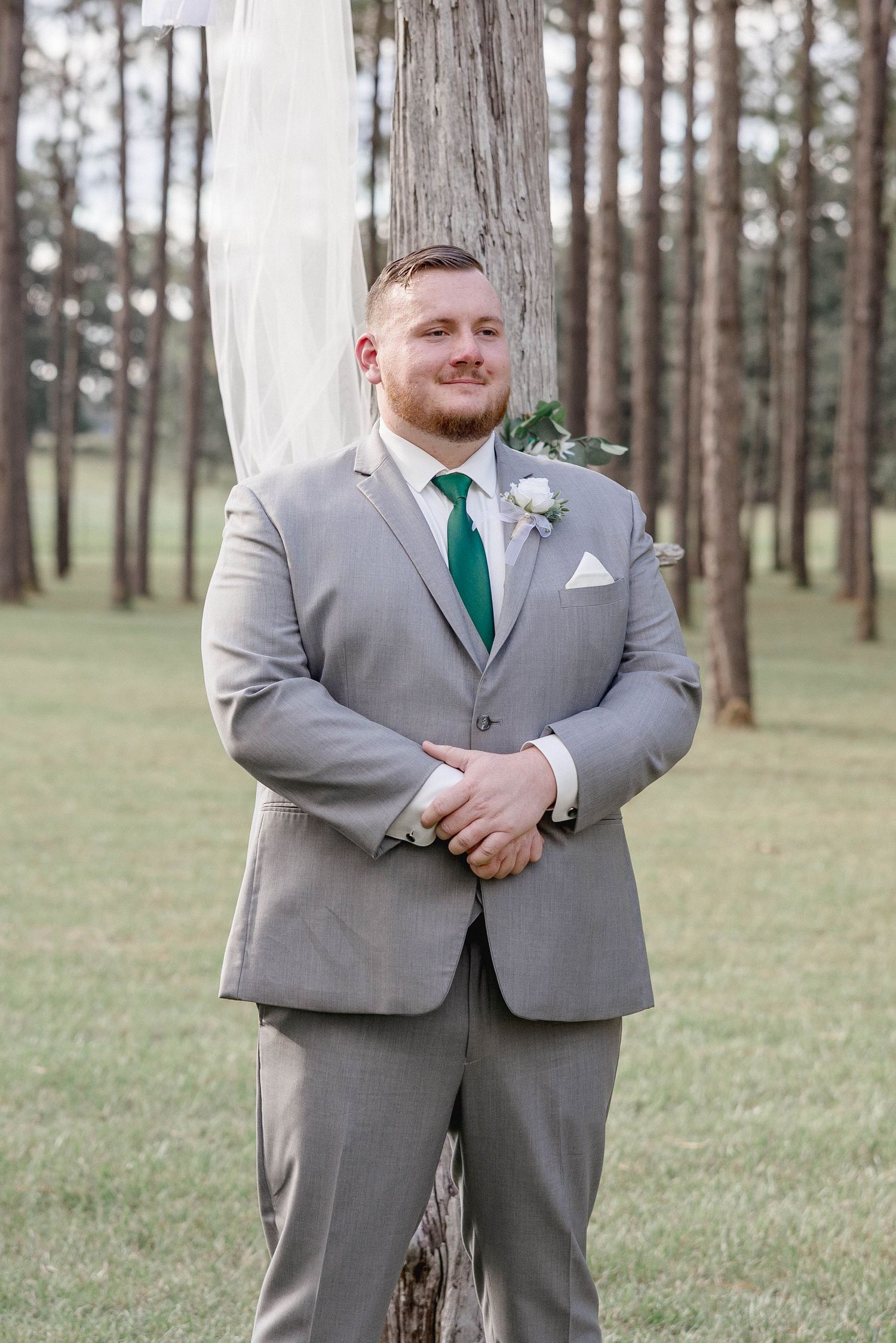 Groom in gray suit with green tie and boutonniere standing outdoors at a pine tree wedding ceremony.