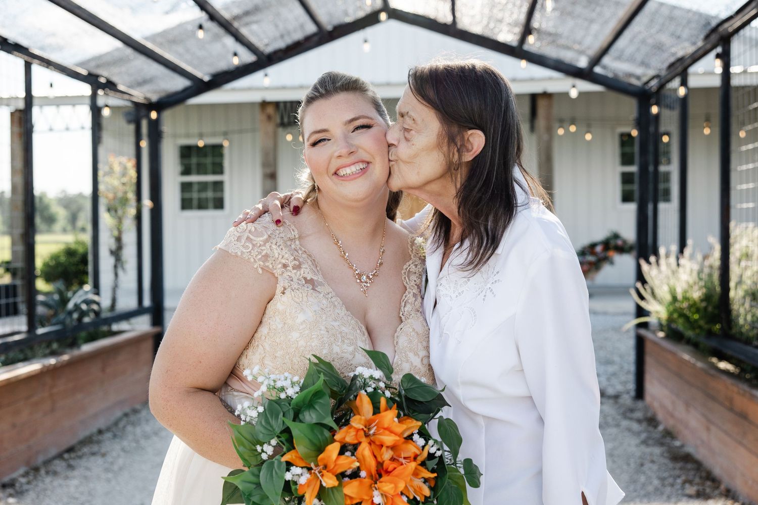 Older woman kisses smiling bride on cheek while bride holds vibrant orange flower bouquet outdoors.