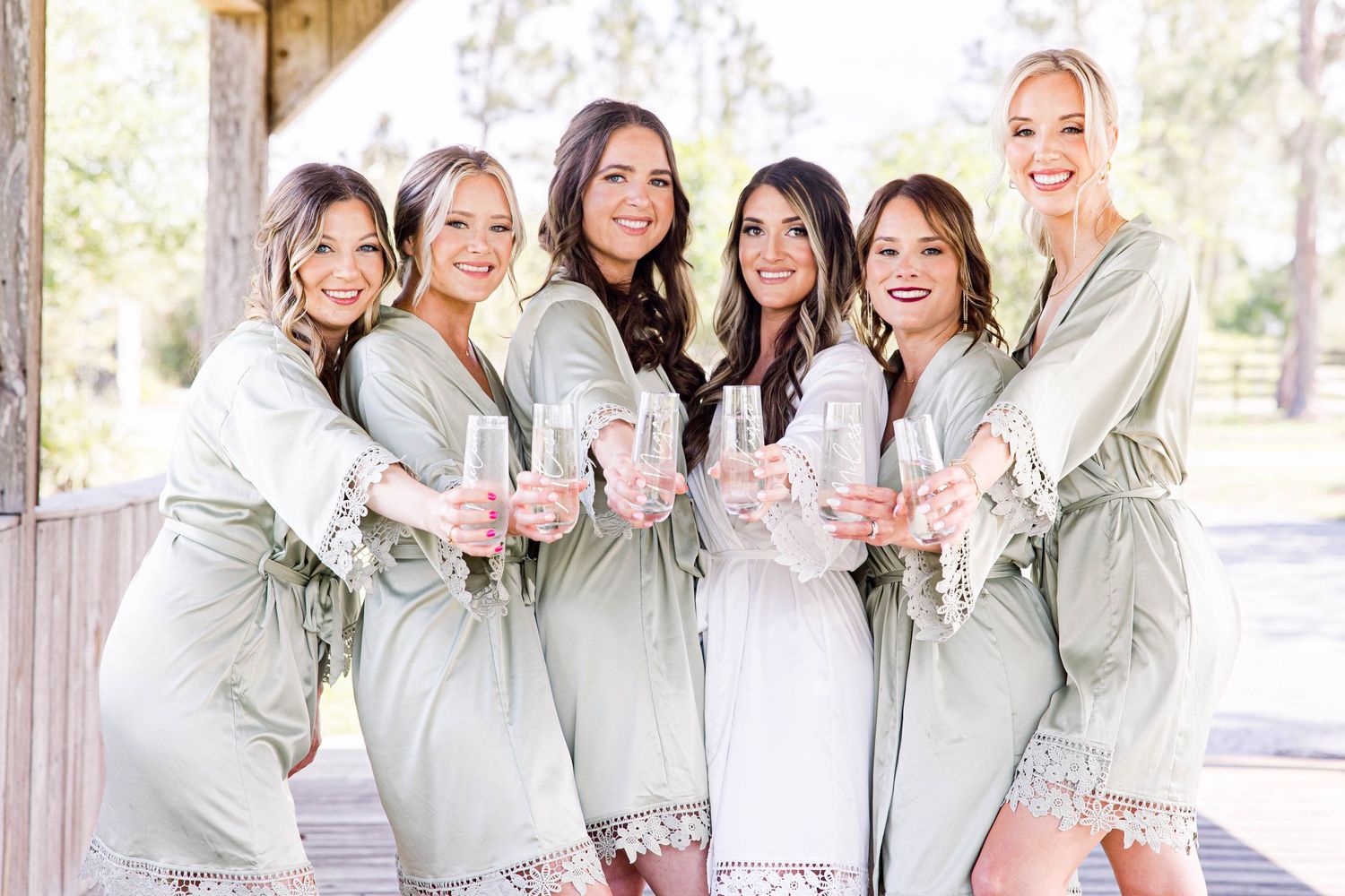 Six bridesmaids in sage green robes toasting with champagne glasses at a rustic wedding venue.