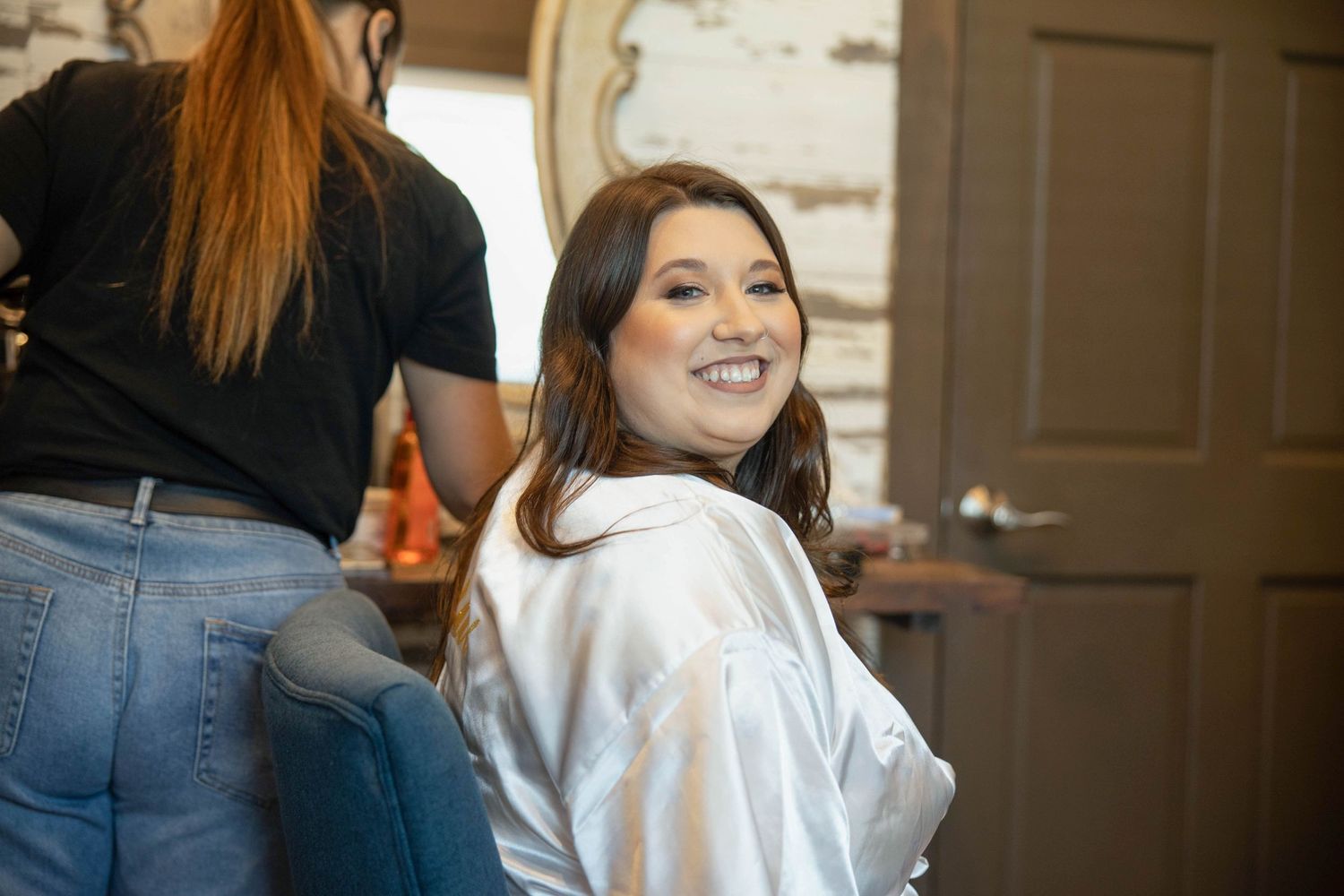 Smiling bride in white robe getting hair and makeup done during wedding morning preparations.