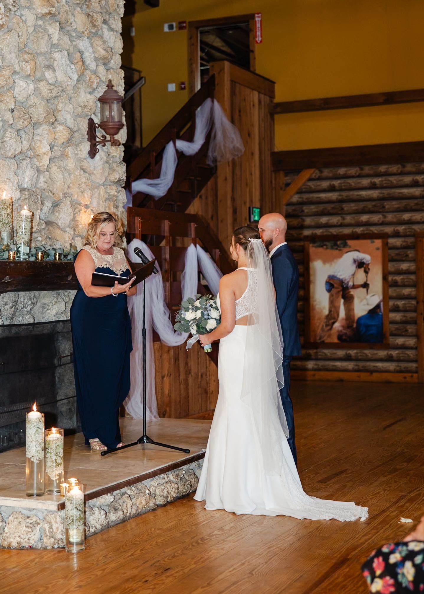 Wedding ceremony inside rustic log cabin venue with stone fireplace, candles, and officiant reading vows.