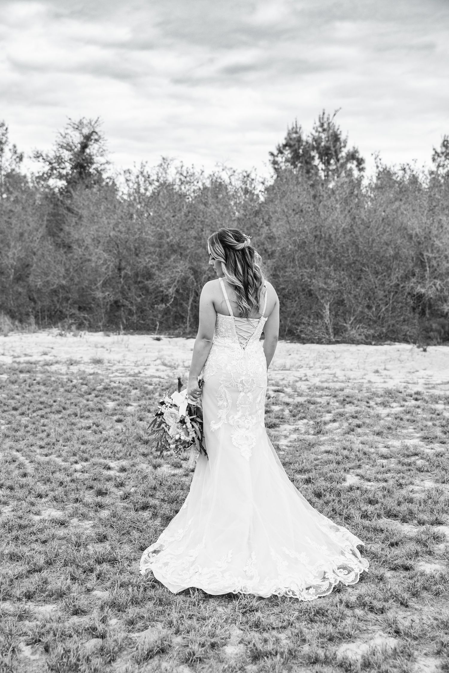 Black and white back view of bride in elegant lace wedding gown holding bouquet in an open field.