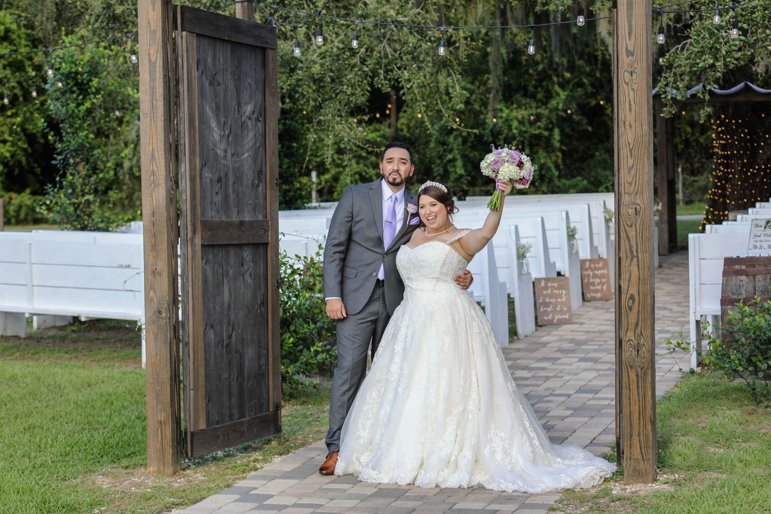 Bride and groom celebrate at rustic outdoor wedding venue entrance with wooden doors and white chairs.