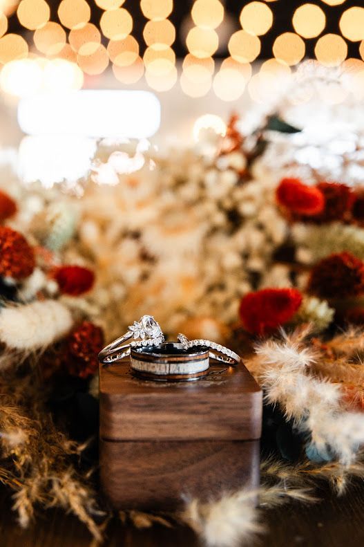 Wedding rings displayed on wooden box surrounded by dried floral arrangement and bokeh lights.