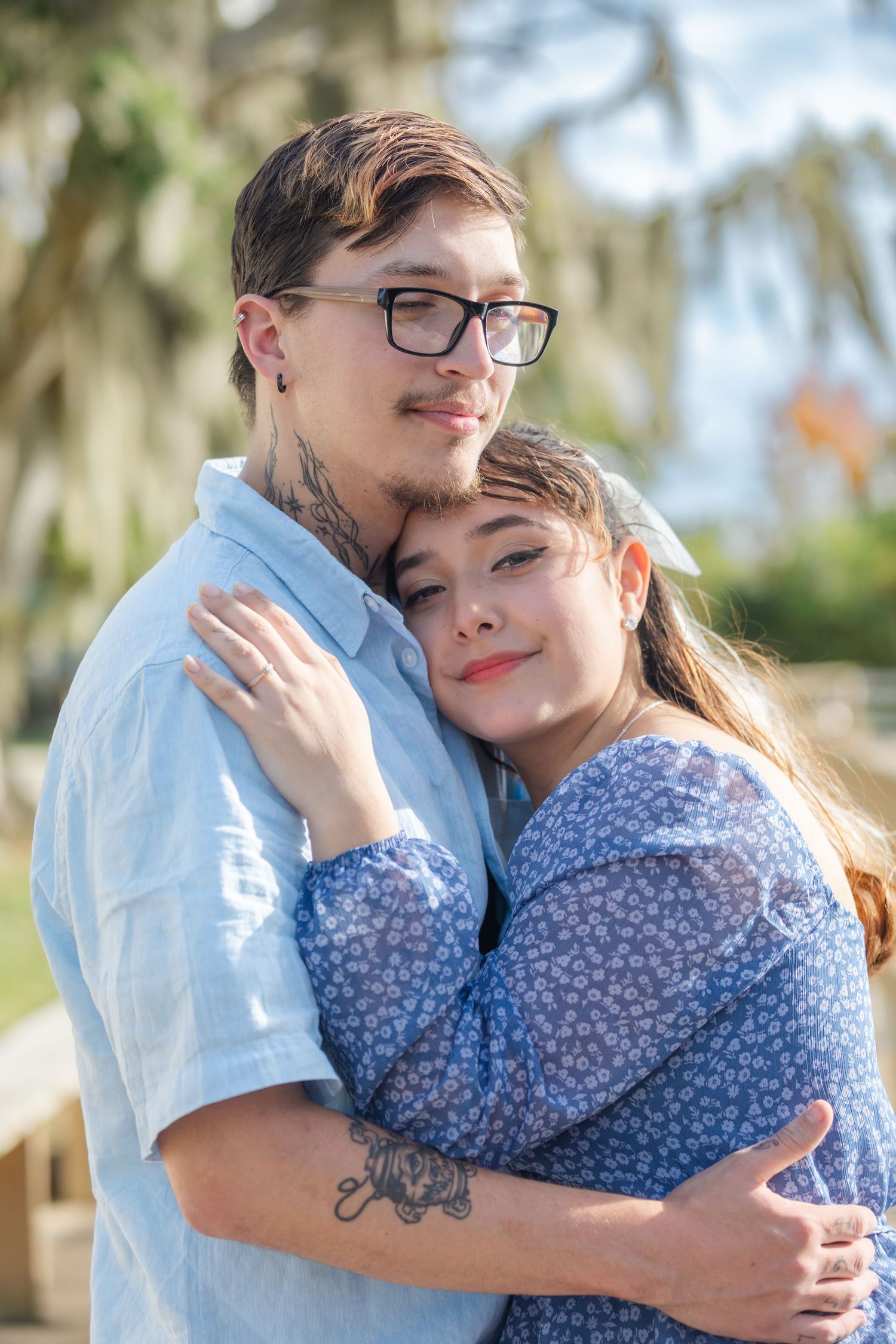 Young tattooed couple in casual blue outfits share a tender embrace outdoors under Spanish moss.