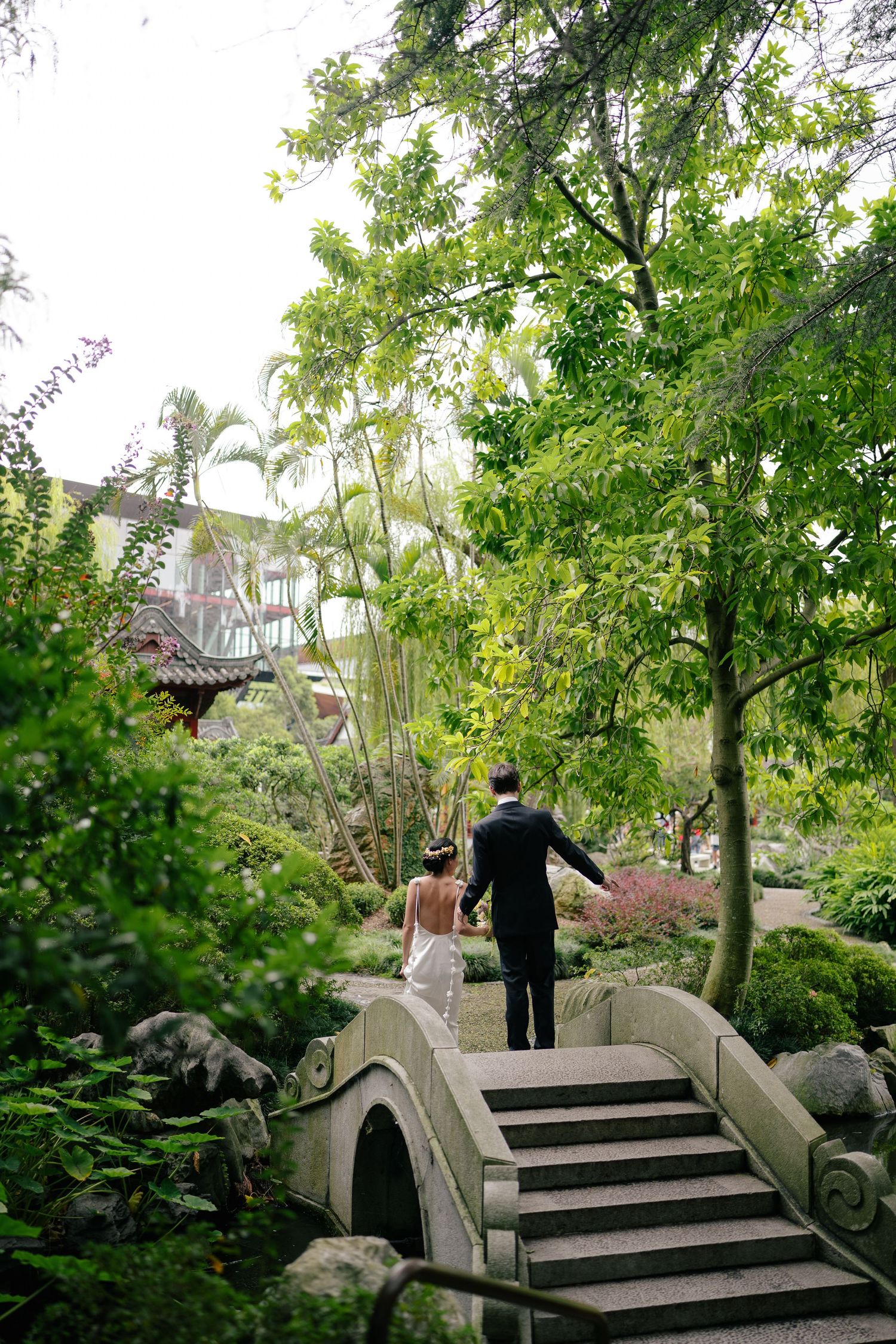 Bride and groom walk hand-in-hand up stone steps in a lush garden with Asian-inspired architecture.