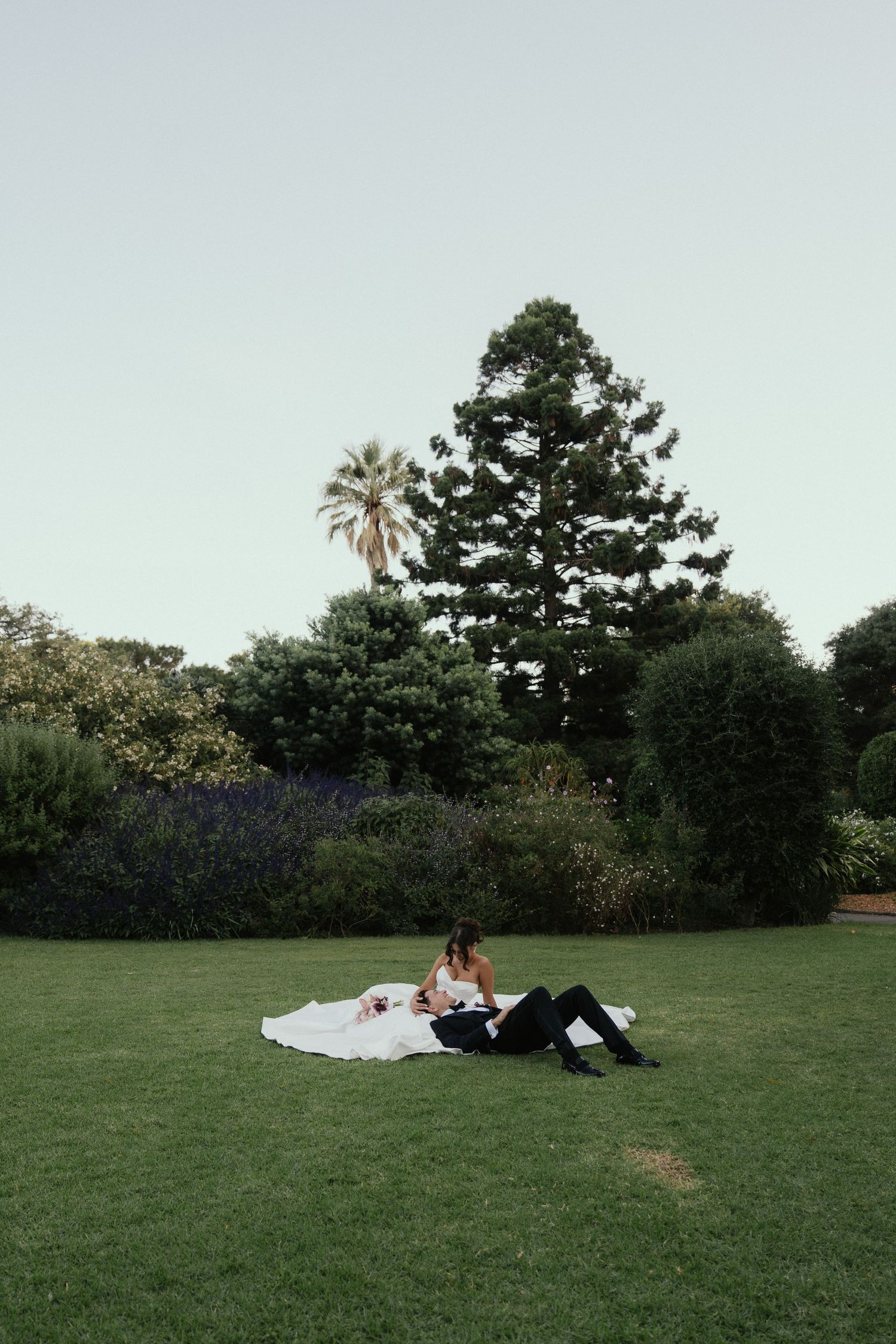Bride and groom relaxing on lush green lawn surrounded by tall trees and colorful garden at dusk.