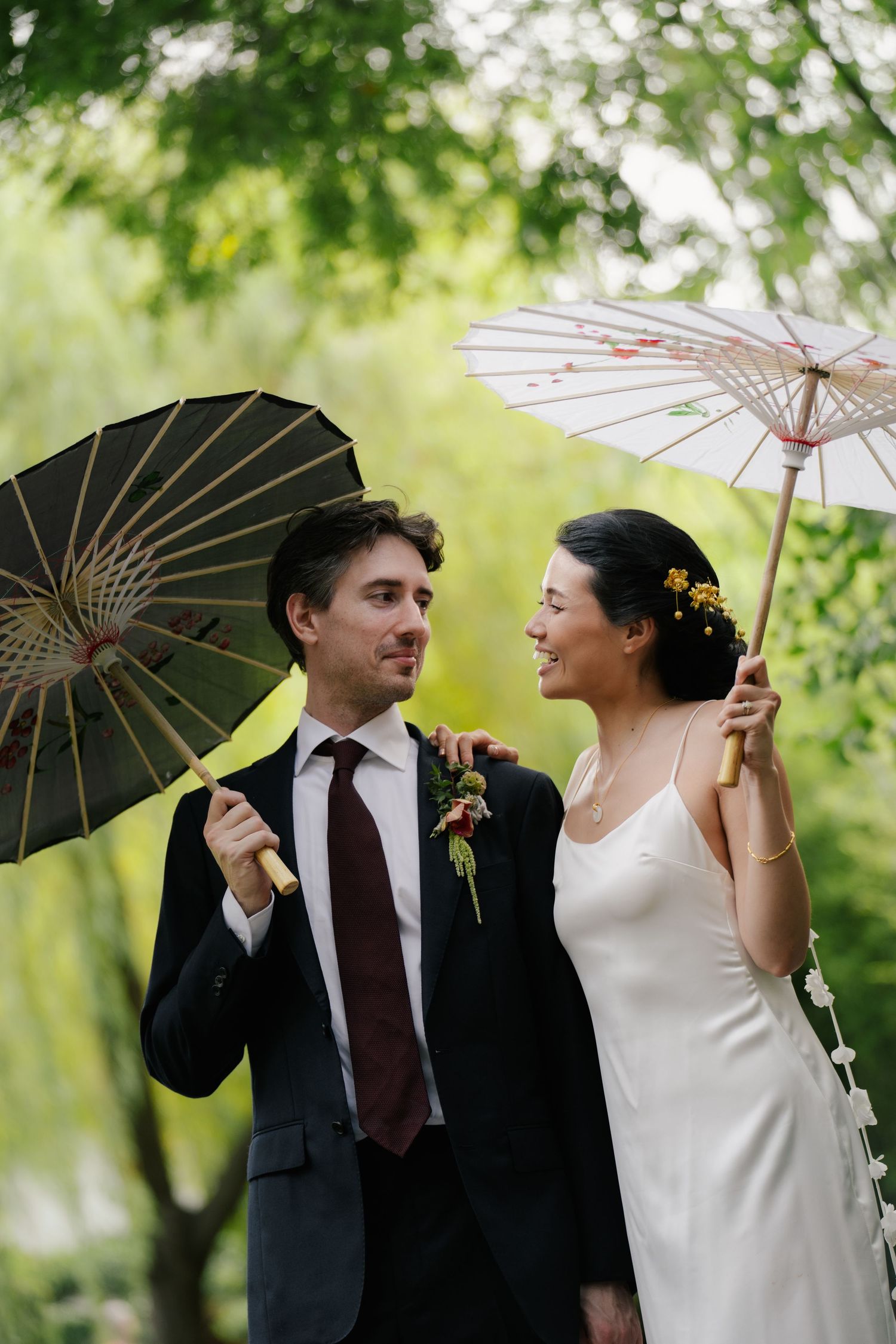 Bride and groom smiling at each other holding decorative parasols outdoors among lush green trees on their wedding day.