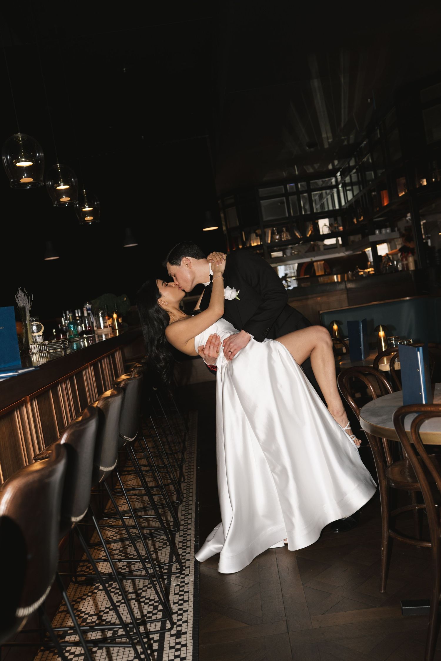 Groom dips bride in elegant white gown for a romantic kiss inside a dimly lit upscale bar venue.