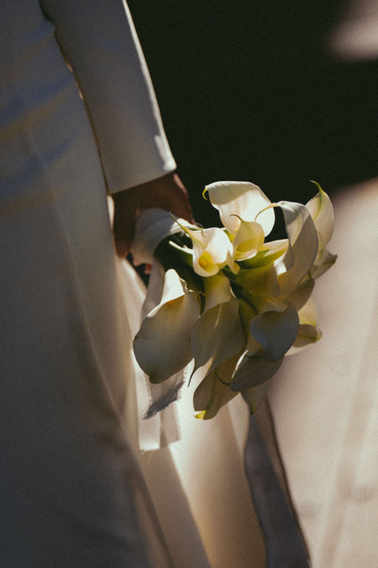 Person in white suit holding elegant calla lily wedding bouquet tied with ribbon in soft natural light.