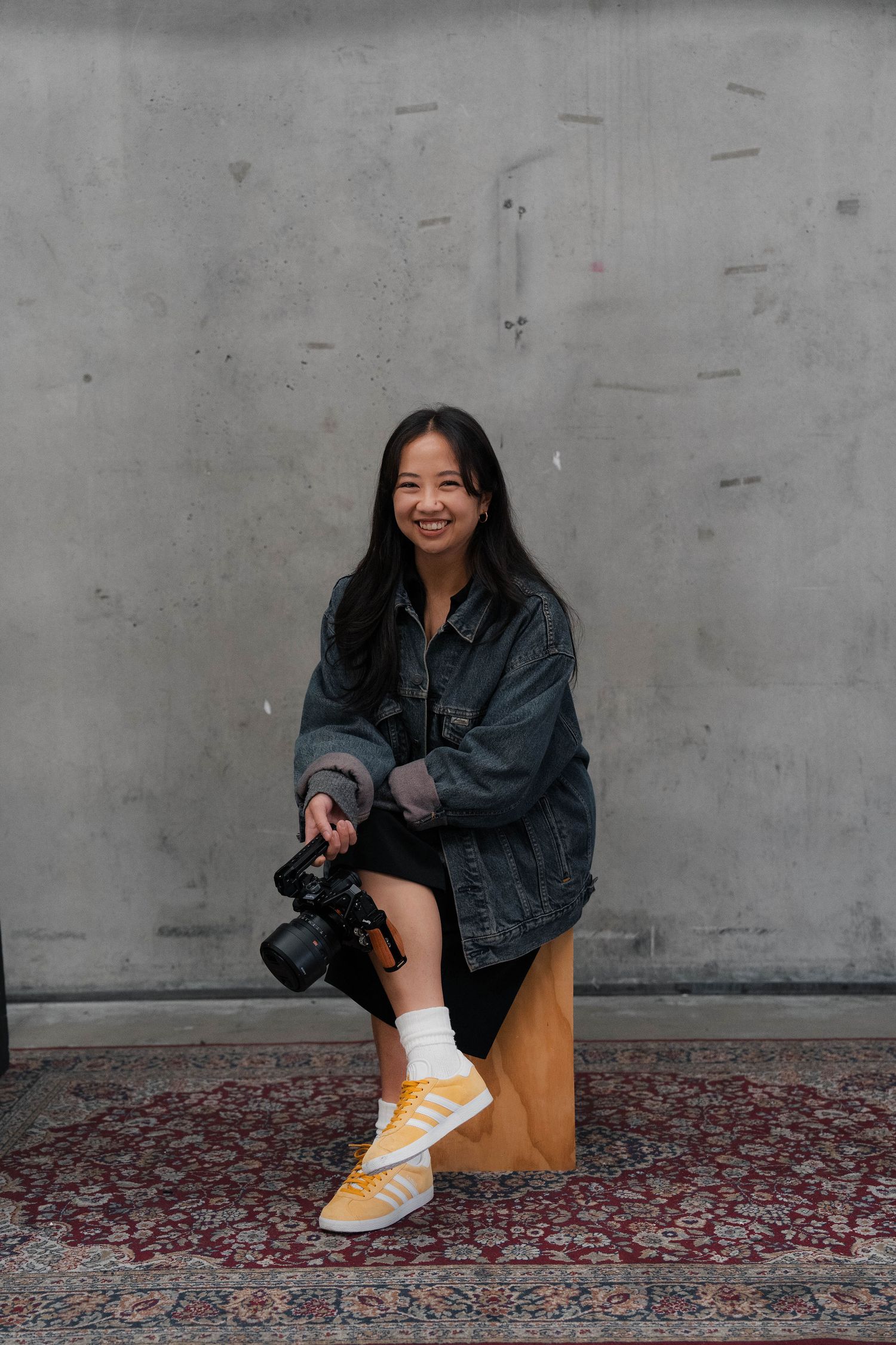 Young woman in denim jacket holding a camera, smiling while seated on a stool against a gray wall.