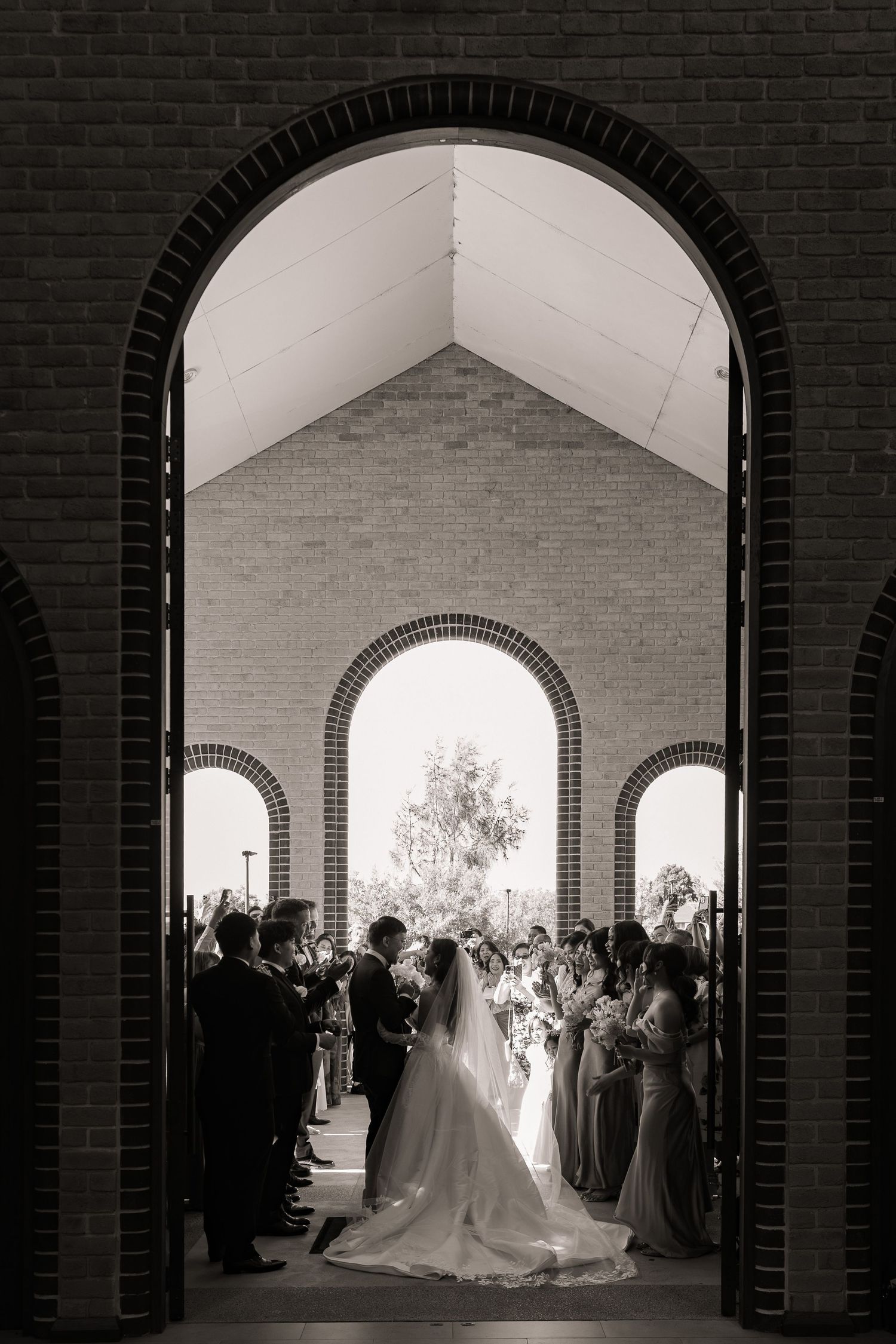 Black and white photo of bride and groom kissing at church altar surrounded by wedding party through arched doorway.