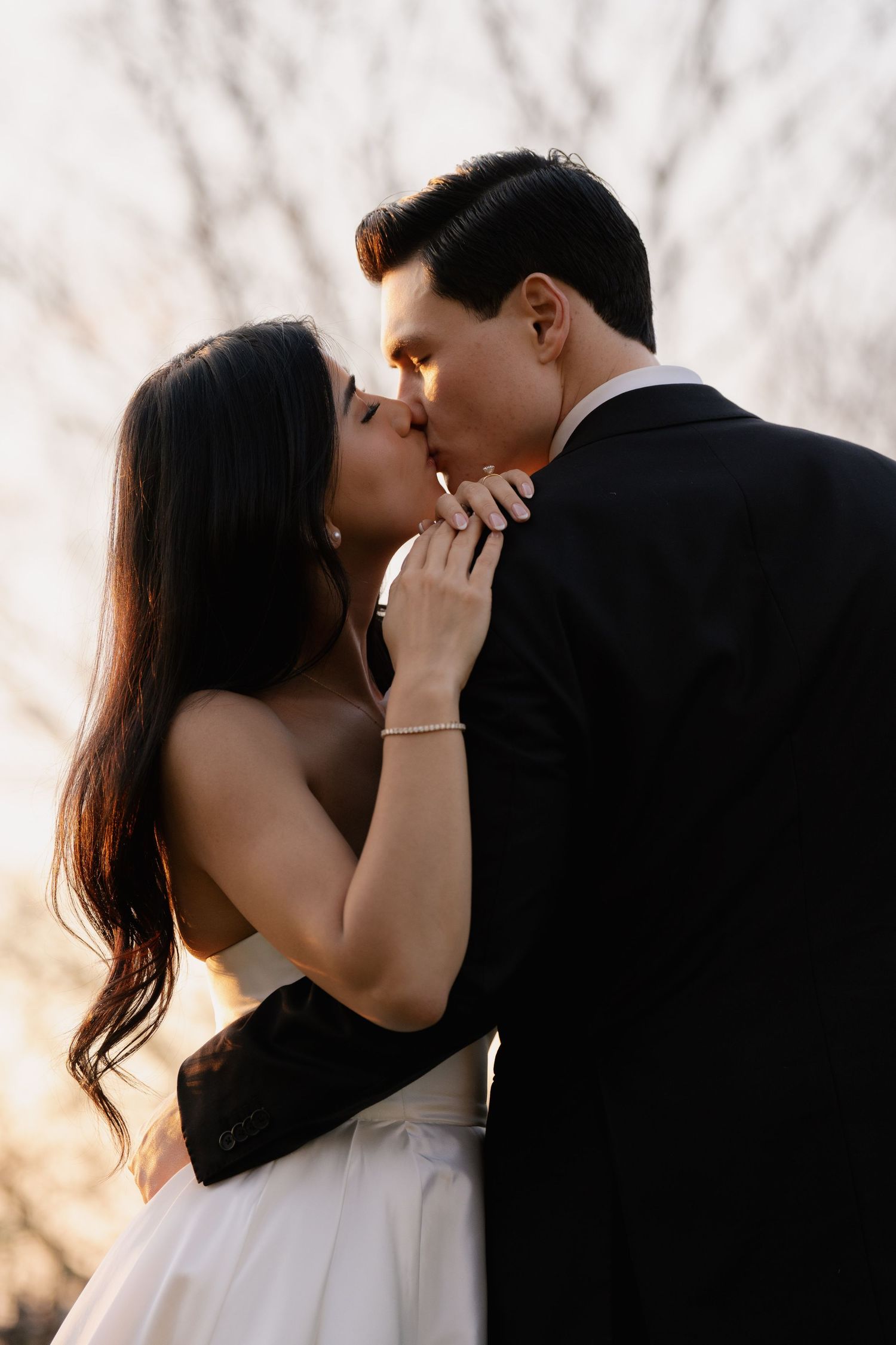 A bride and groom share a romantic kiss outdoors at sunset, with bare winter trees in the soft background.