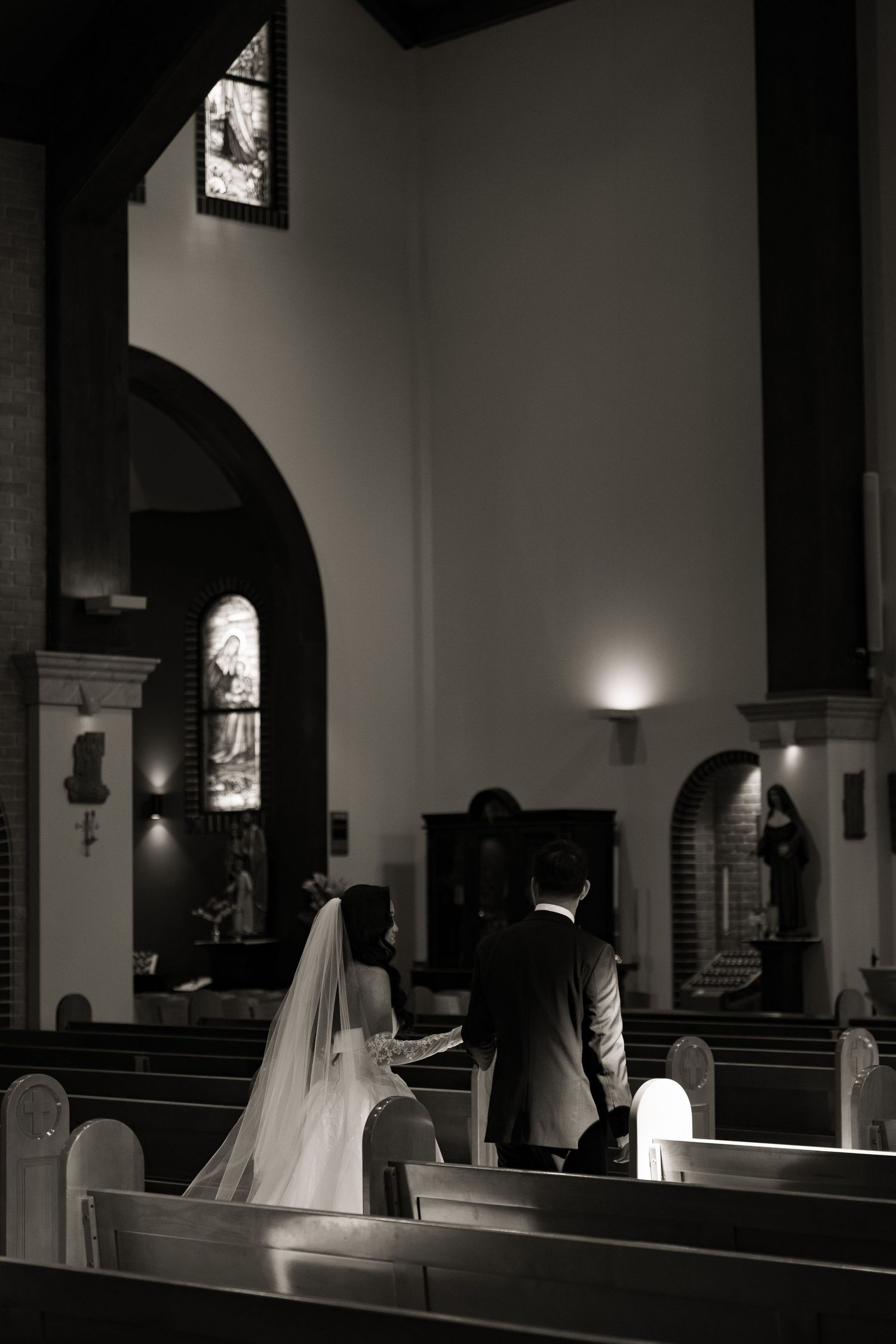 Black and white photo of bride and groom kneeling at church altar during wedding ceremony with stained glass windows.