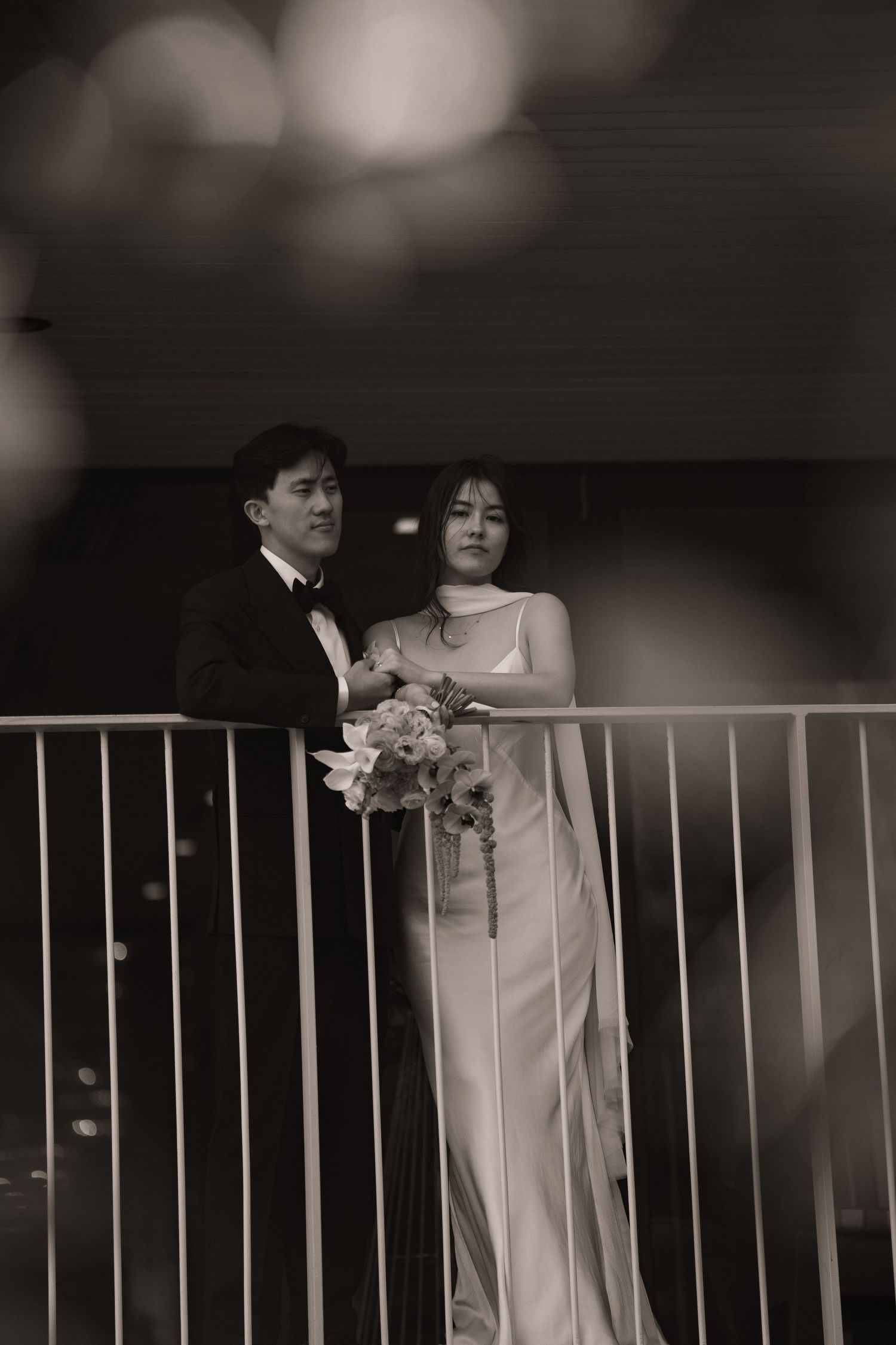 Black and white photo of a bride in a sleek gown holding a bouquet beside a groom in a tuxedo on a balcony.