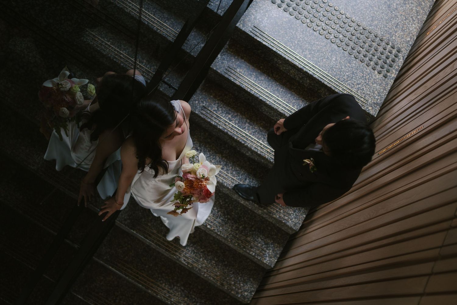 Aerial view of bride holding bouquet and groom standing on stairs in dramatic lighting.