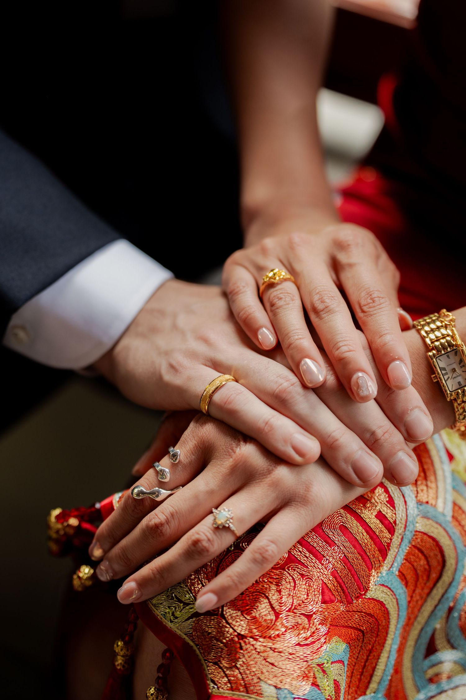 Couple's hands stacked together wearing gold wedding rings and bracelets over a red embroidered Chinese bridal qipao.