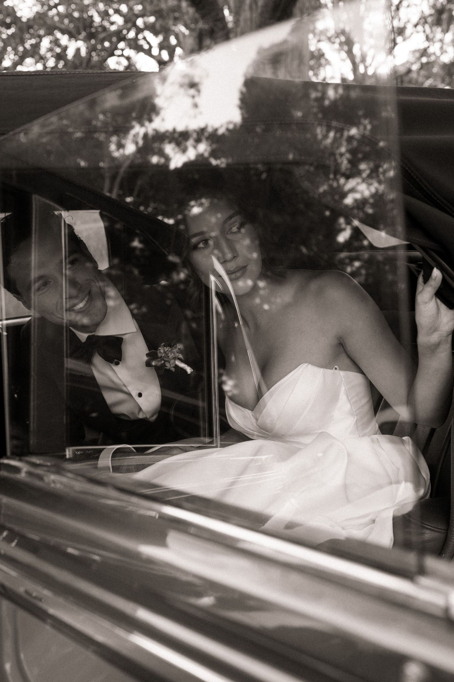 Black and white photo of a smiling bride in white gown and groom in tuxedo sitting together inside a vintage wedding car.