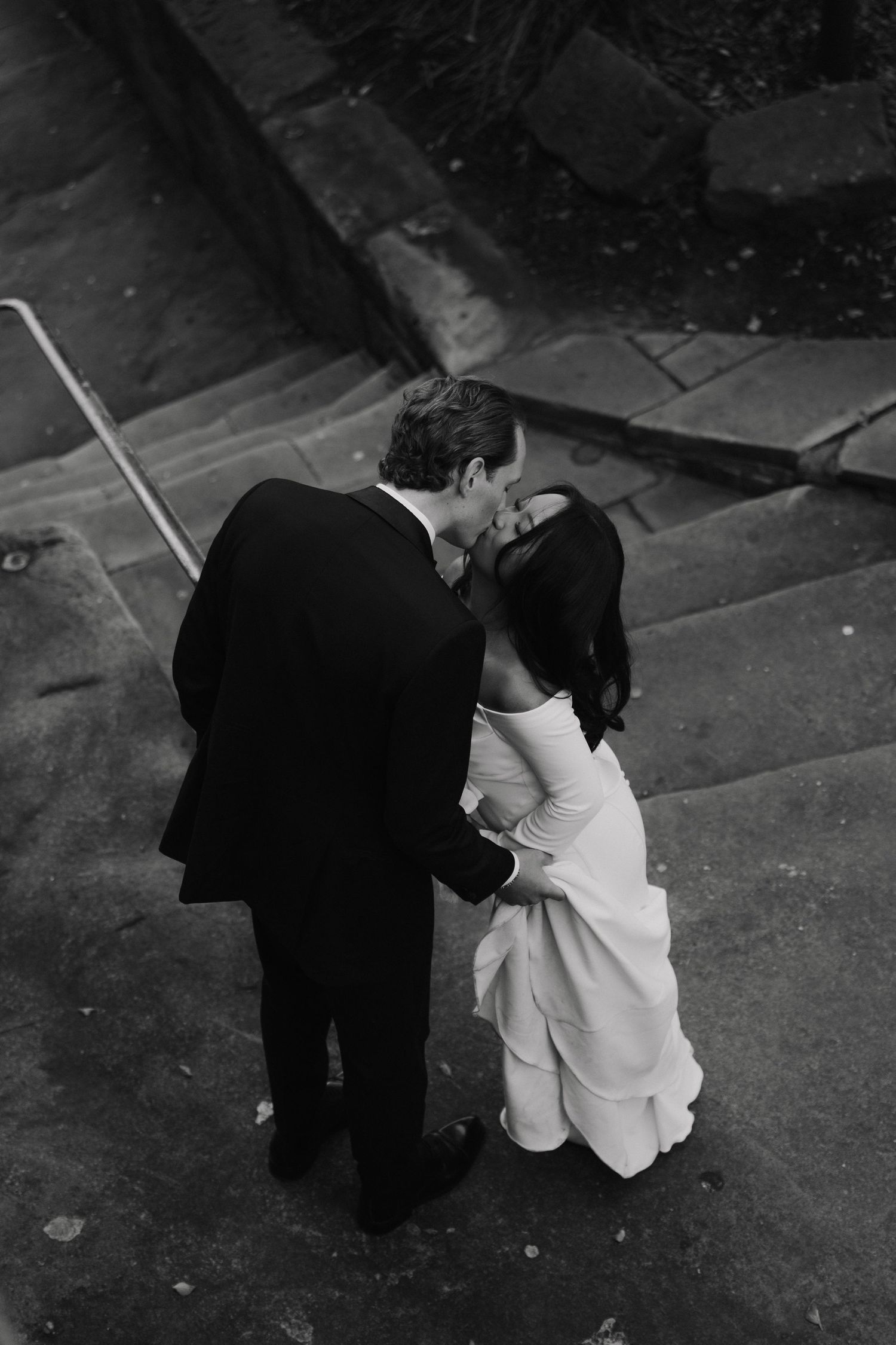 Black and white photo of a bride and groom sharing a kiss on stone steps, captured from above.