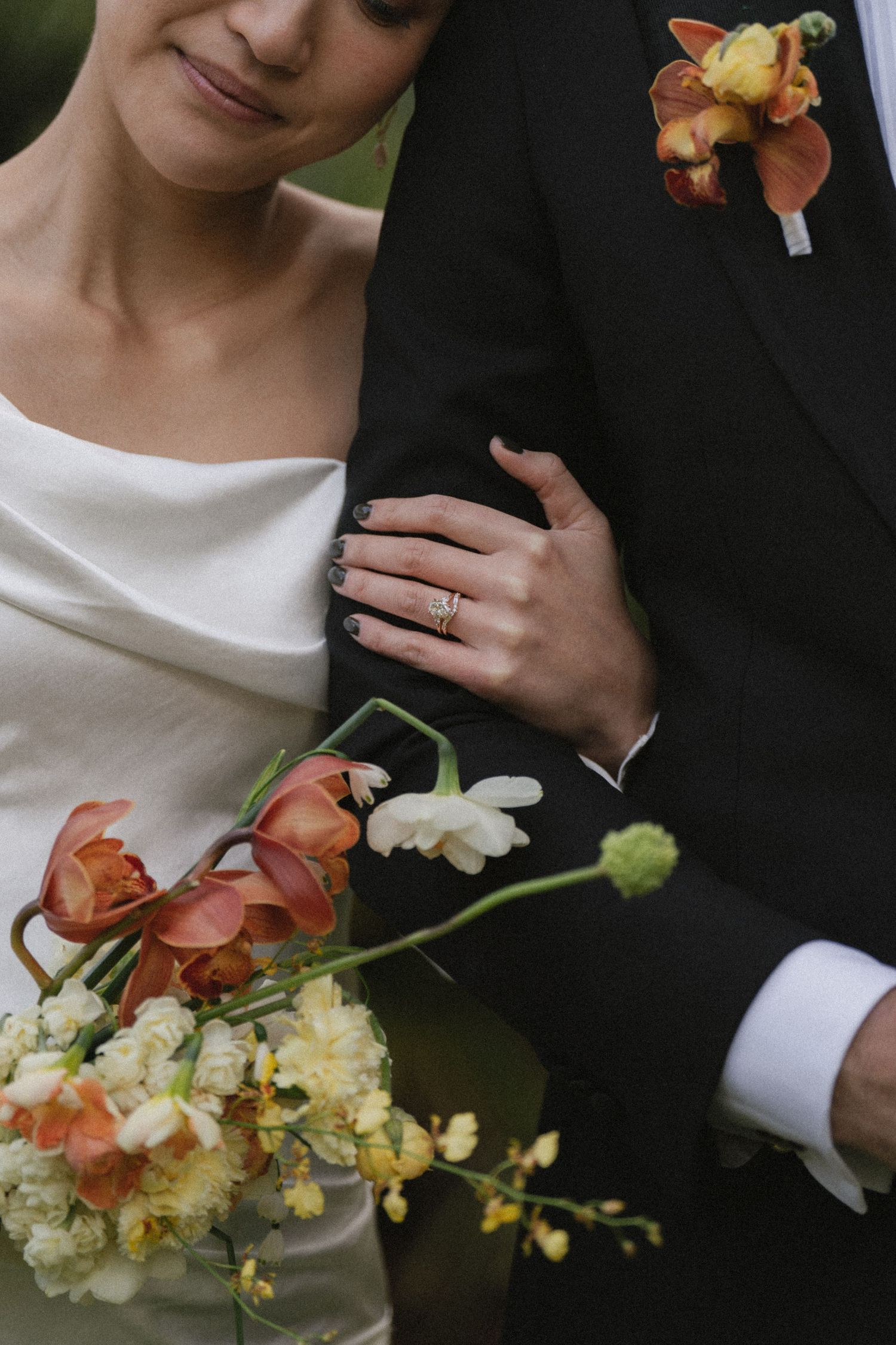 Bride holding orange and white wedding bouquet while resting hand on groom's black suit, showing engagement ring.