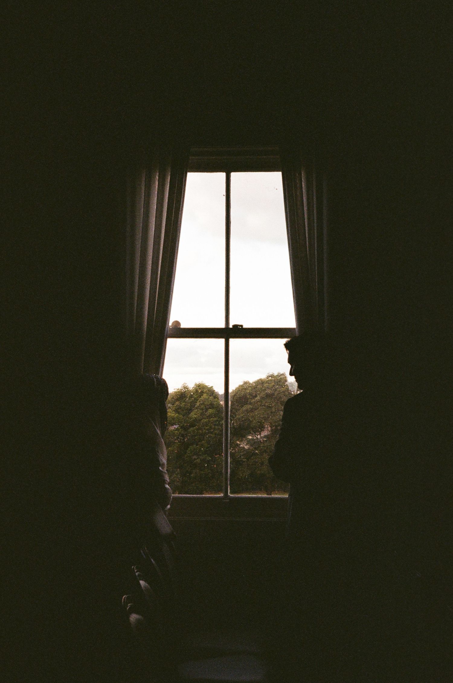 Two silhouetted figures stand before a bright window with curtains, overlooking lush green trees in a dark room.