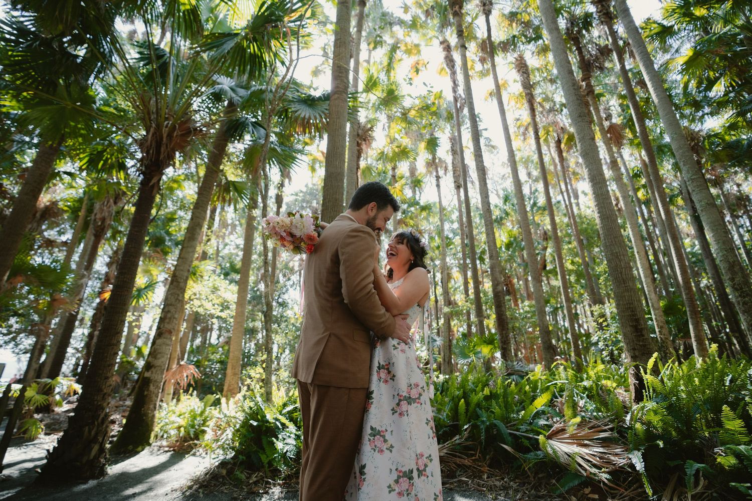 Couple dancing romantically among tall palm trees in a lush tropical garden, groom in tan suit, bride in floral dress.