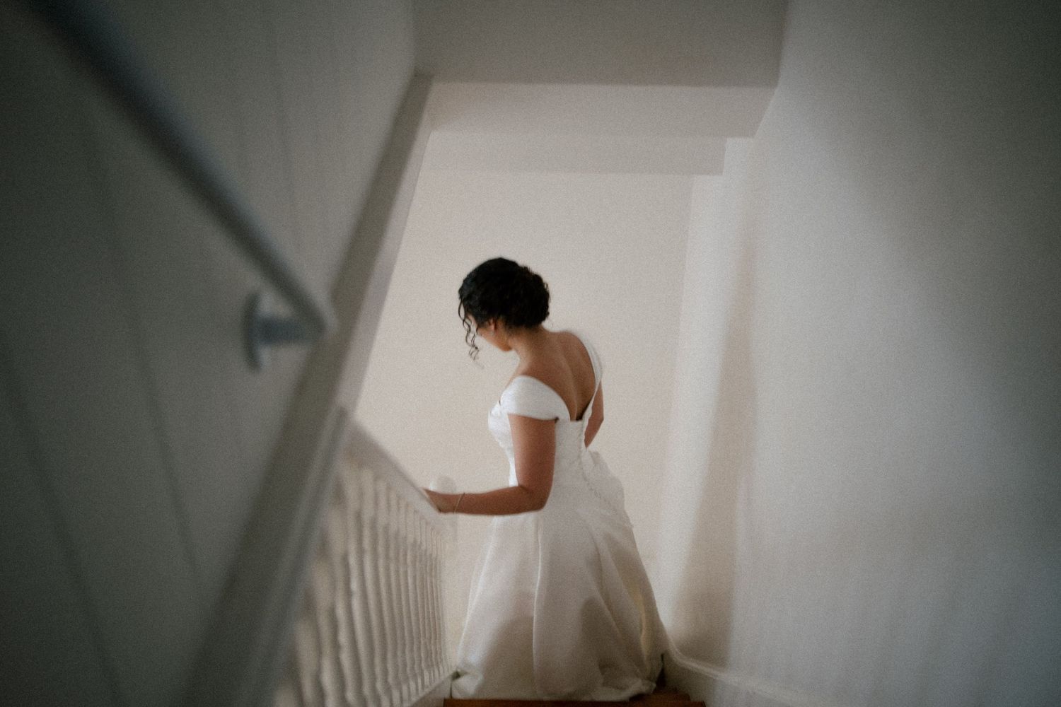 Bride in elegant white backless wedding gown ascending staircase, viewed from behind in soft natural light.