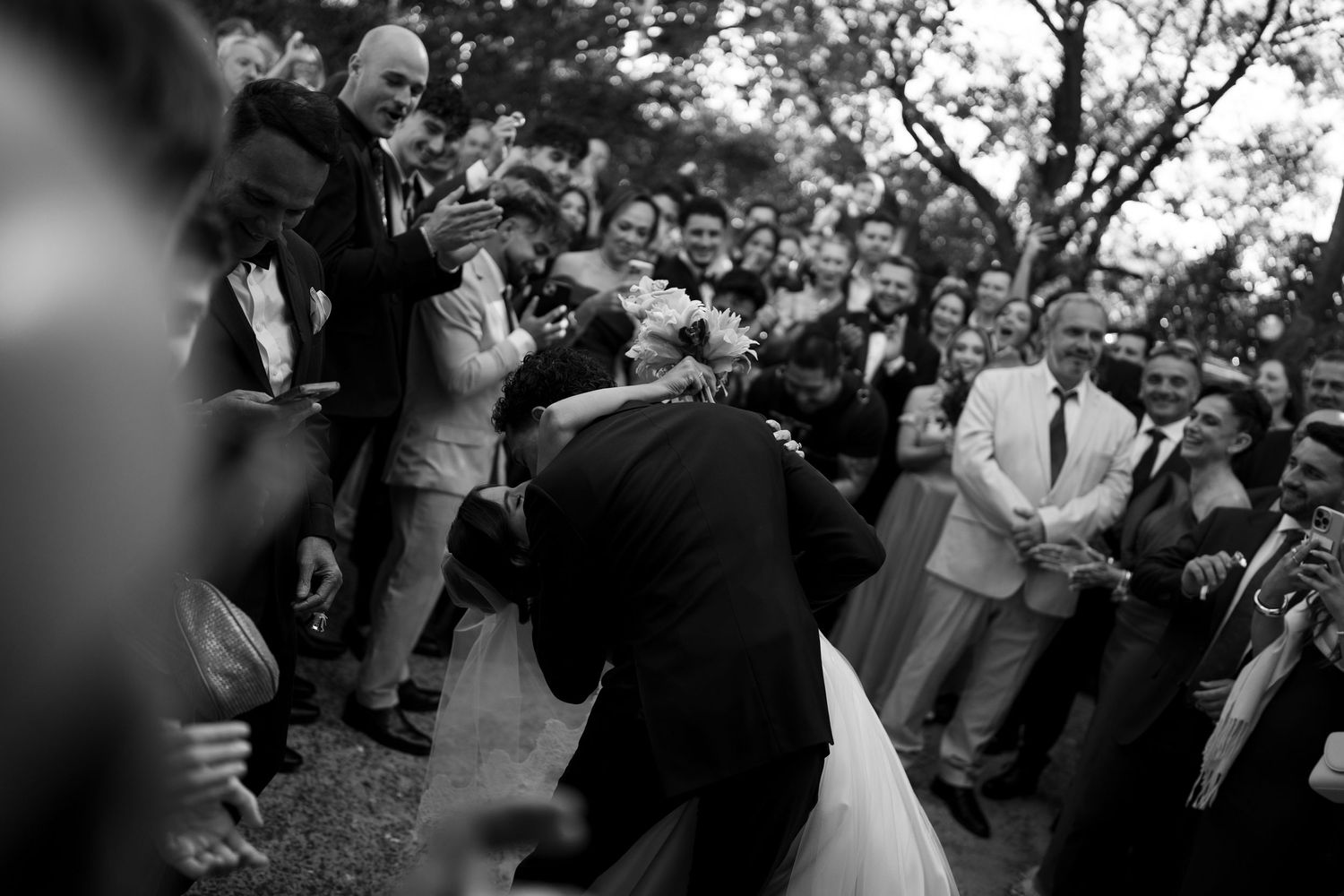 Black and white wedding photo of couple sharing a kiss surrounded by cheering guests outdoors.