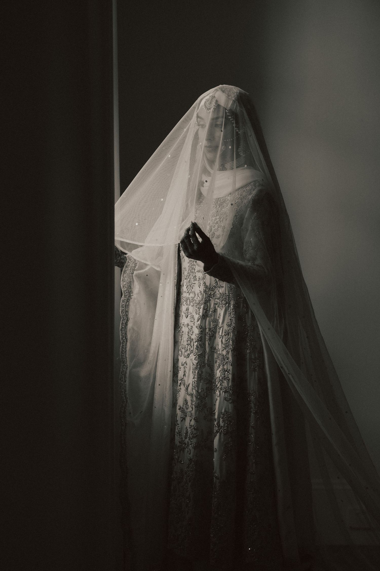 Vintage black and white photograph of a bride wearing a delicate lace wedding dress and veil in dramatic lighting.