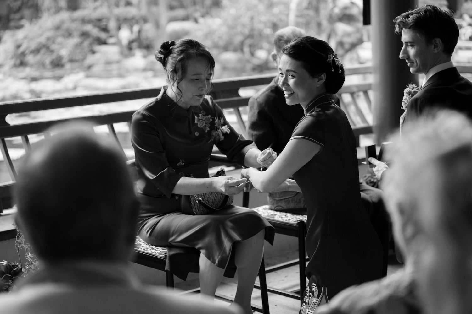 Black and white photo of two women holding hands at a wedding ceremony, seated on a bench with guests nearby.