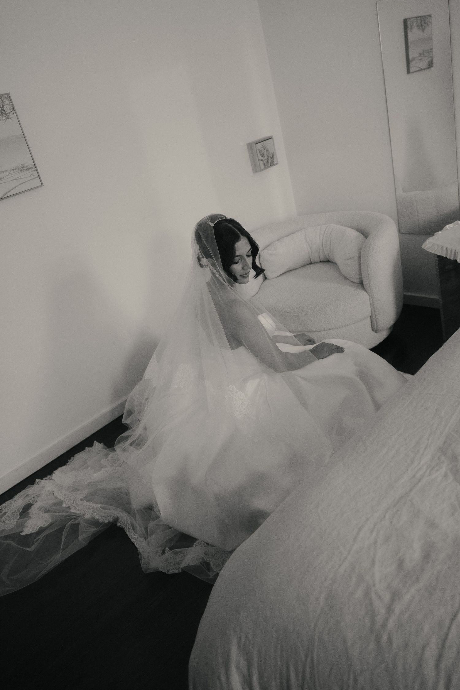Black and white photo of a bride in a flowing wedding gown and veil, seated in a chair reading a letter in a hotel room.