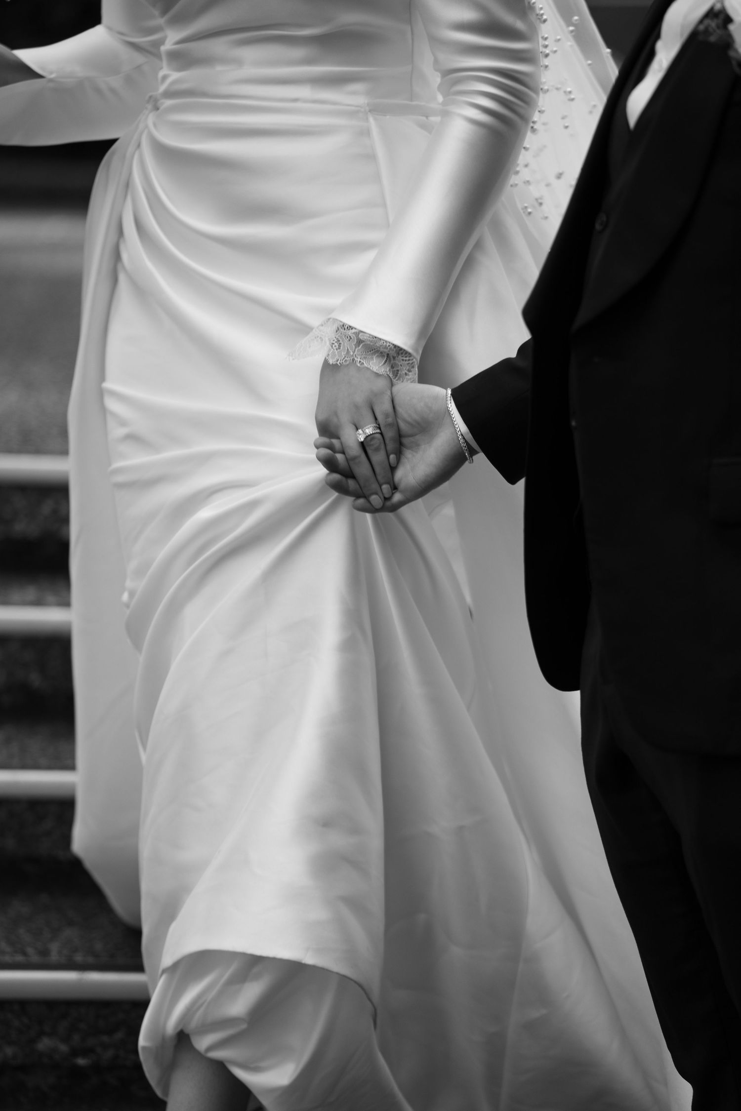 Black and white photo of bride and groom holding hands, showcasing wedding ring and elegant white bridal gown.