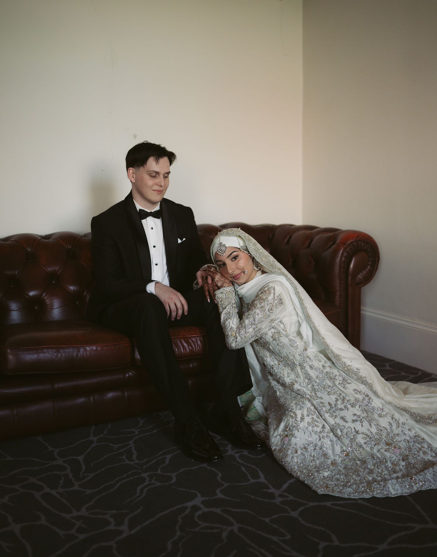 Bride in white lace hijab gown leans on groom in black tuxedo seated on brown leather sofa at wedding.