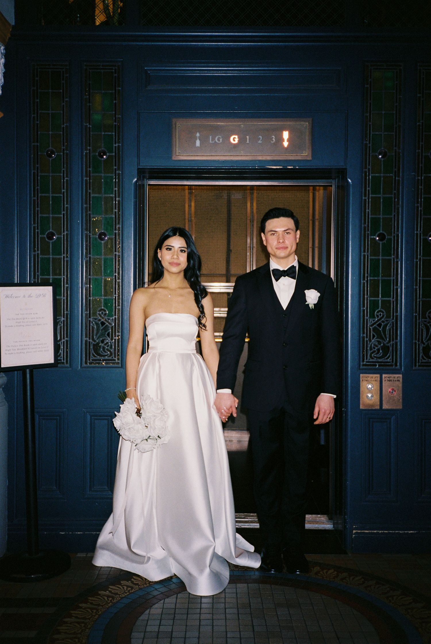 Bride in strapless white satin gown and groom in black tuxedo hold hands standing before ornate blue elevator doors.