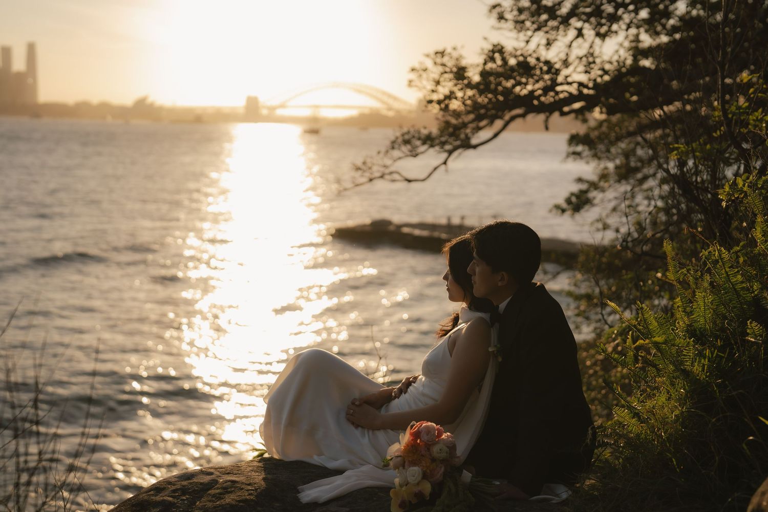 Bride and groom sit together at sunset by the water with a bridge glowing in the golden light behind them.
