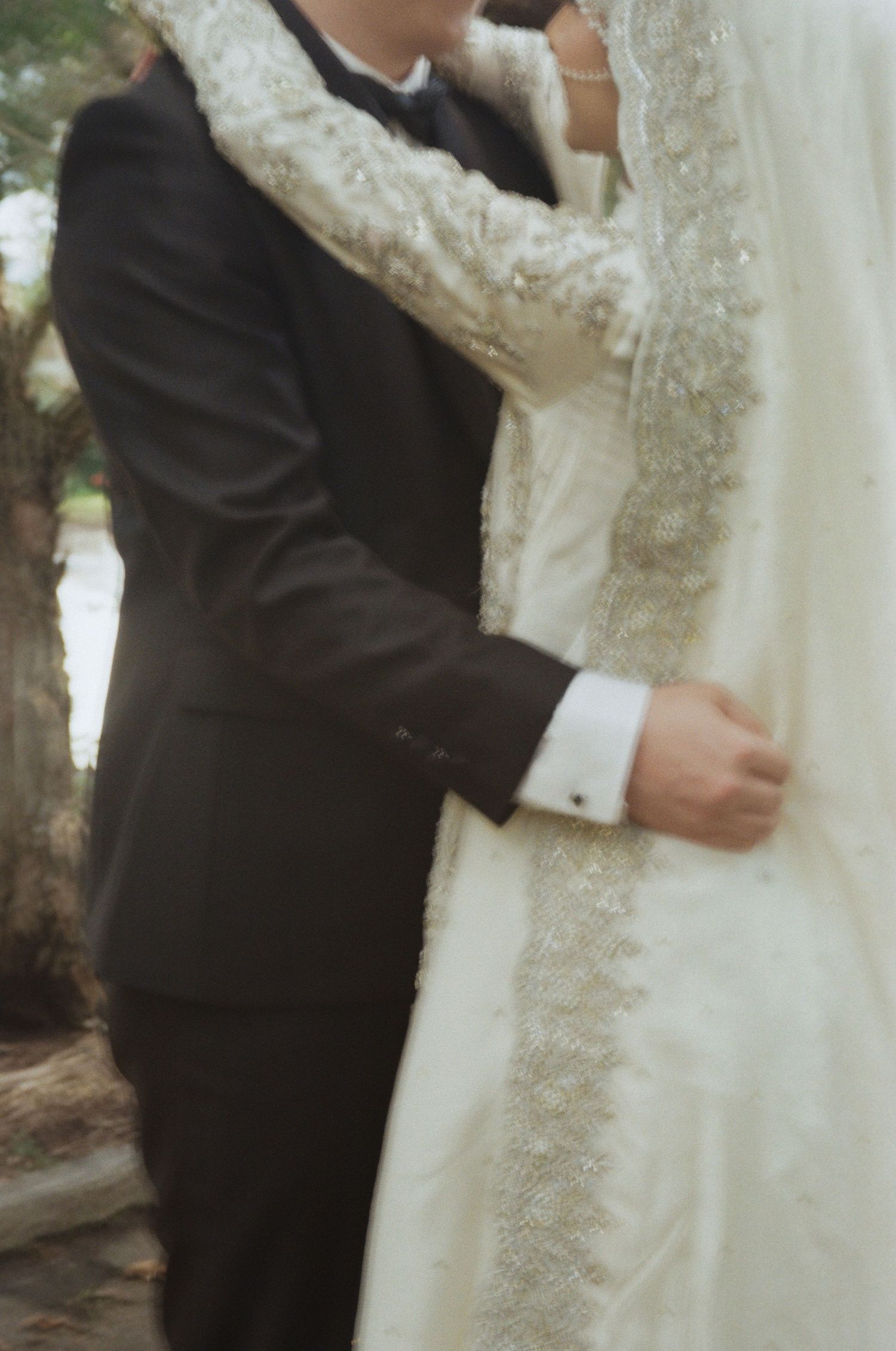A bride in a white lace gown and groom in a black suit embrace outdoors at their wedding ceremony.