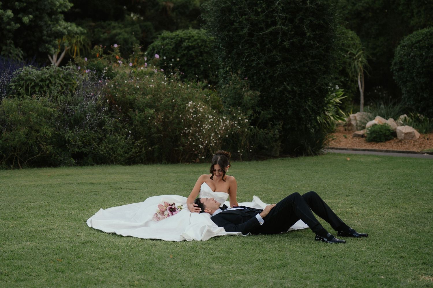 Bride and groom relaxing on green lawn in garden, bride in white gown, groom in black suit, wedding day portrait.