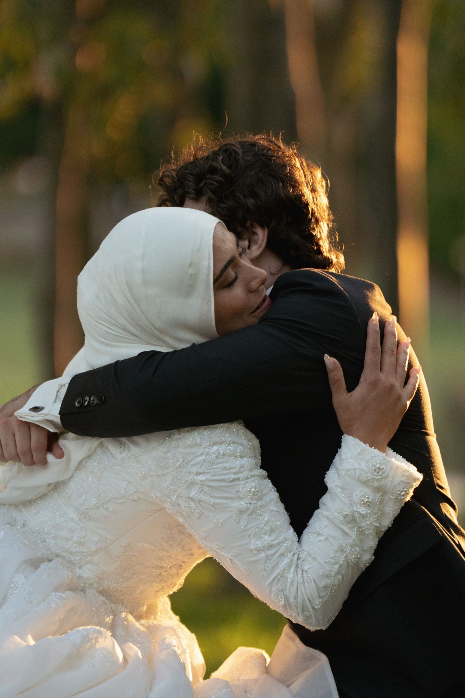 A bride in white hijab and groom in black suit share a tender embrace outdoors at golden hour.