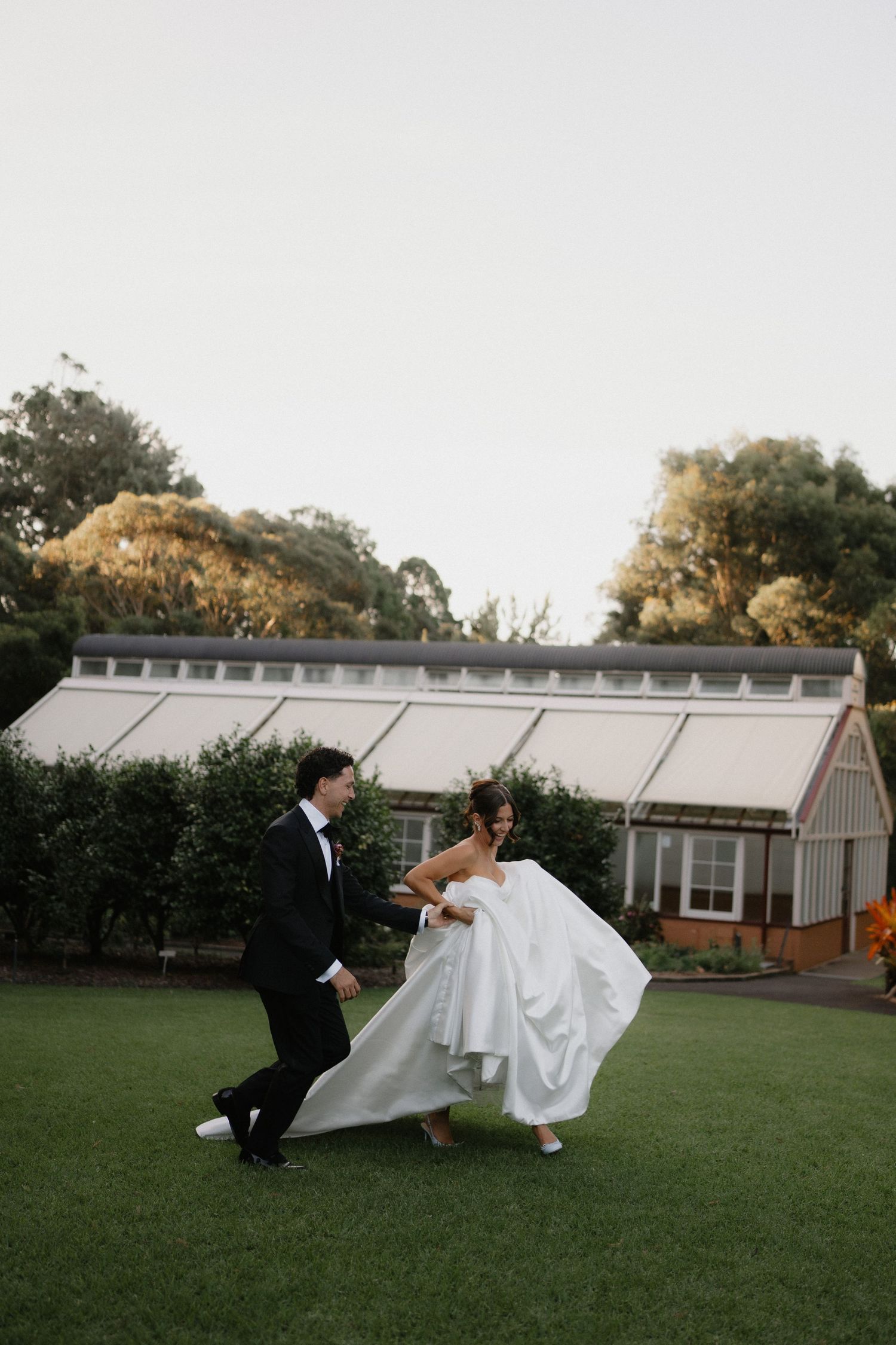 Bride and groom dancing on green lawn outside a greenhouse venue at golden hour wedding celebration.