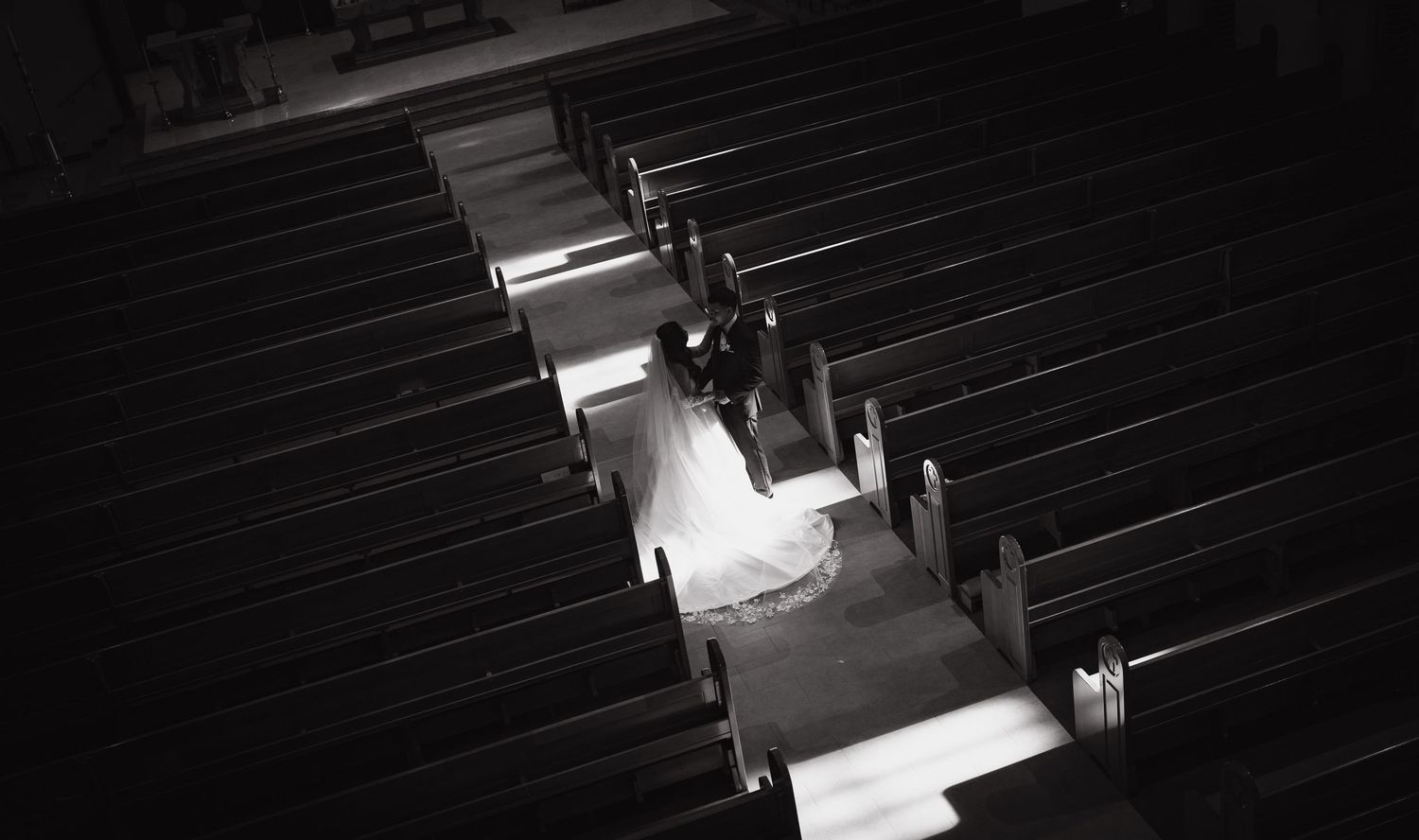 Black and white aerial view of a bride and groom walking down a church aisle surrounded by wooden pews.