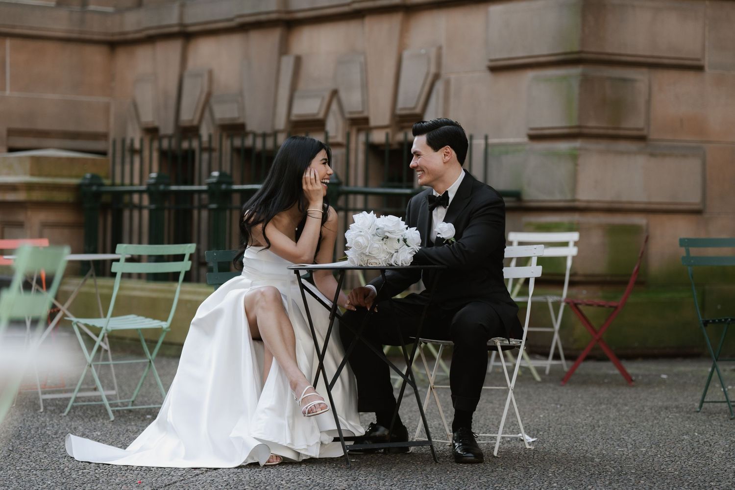 Bride and groom sitting at outdoor cafe table, sharing an intimate moment, bride holding white bouquet in wedding attire.