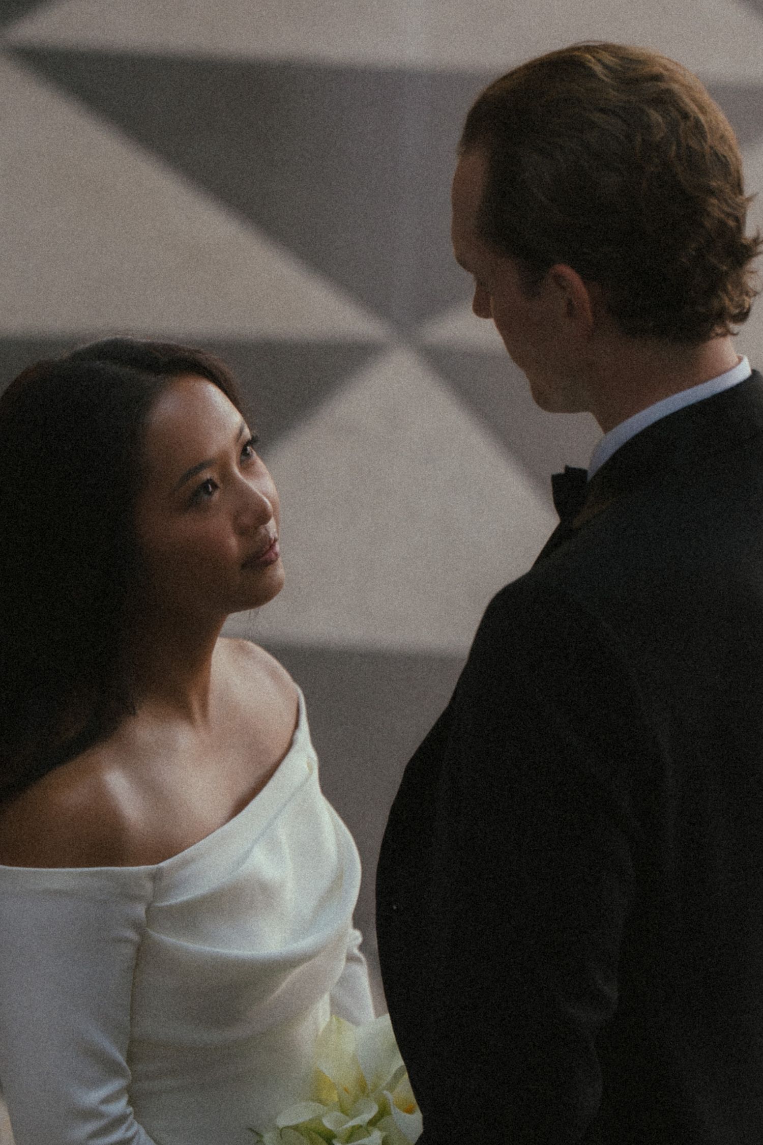 Bride in white off-shoulder gown holds flowers, gazing at groom in black tuxedo during wedding ceremony.