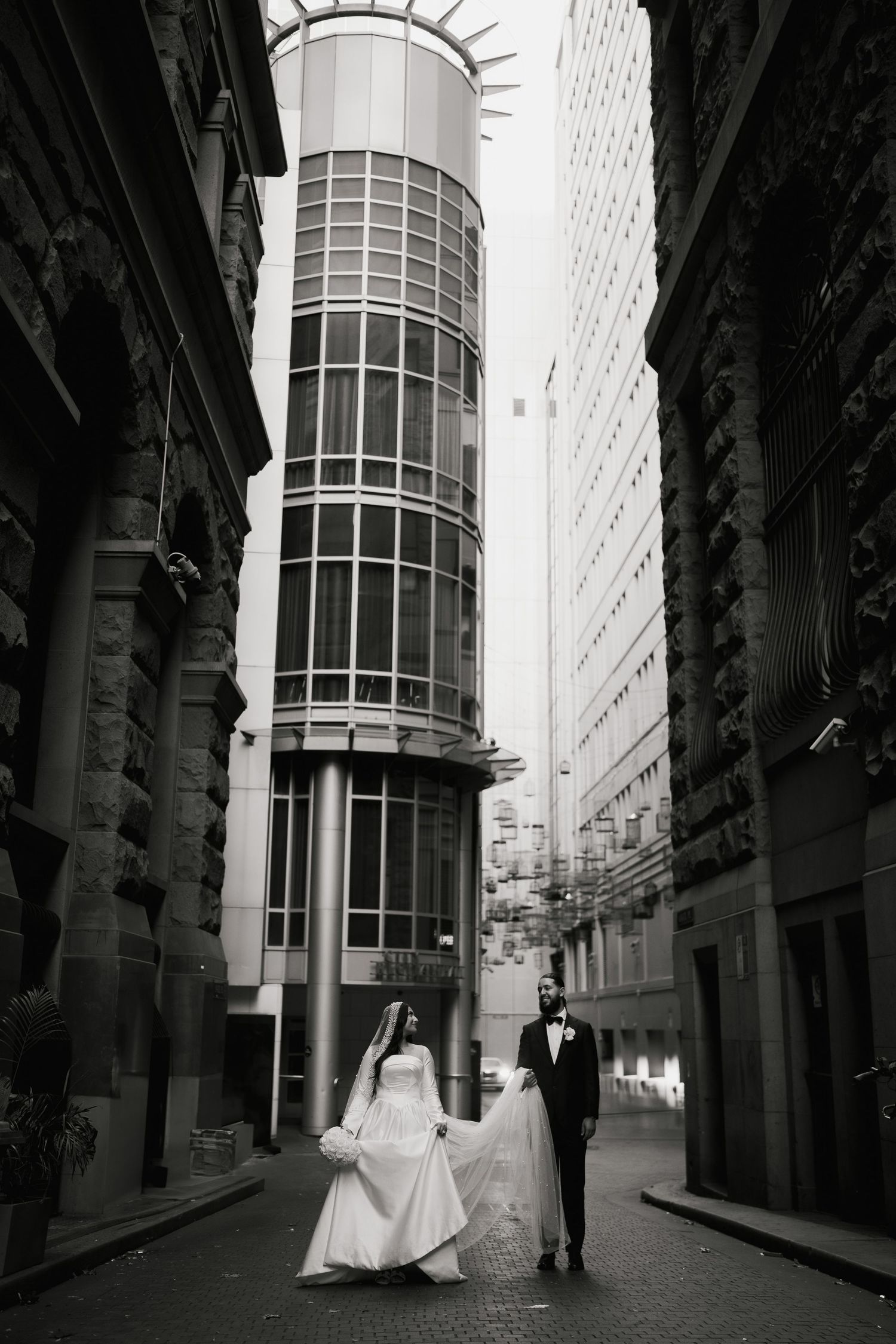 Black and white photo of bride and groom holding hands in urban alley with curved glass building backdrop.