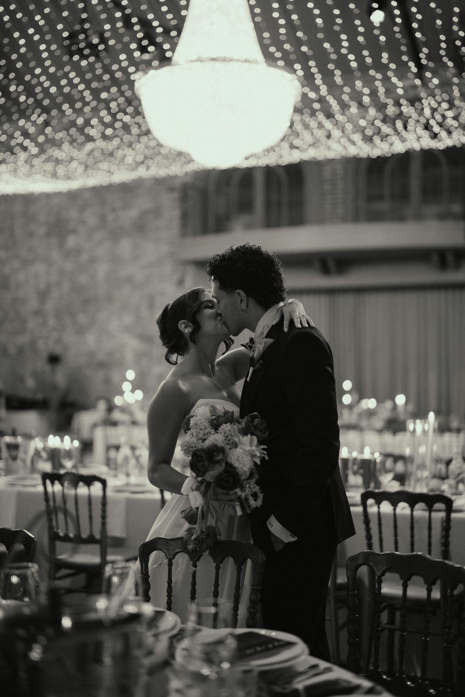 Black and white photo of a bride and groom sharing a kiss under a chandelier at their elegant wedding reception.